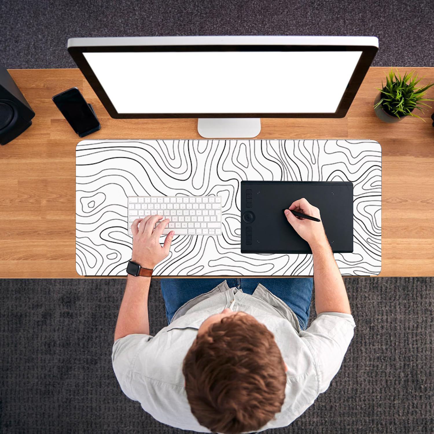 Personne utilise clavier et tablette graphique. Moniteur, téléphone et plante sur bureau en bois.