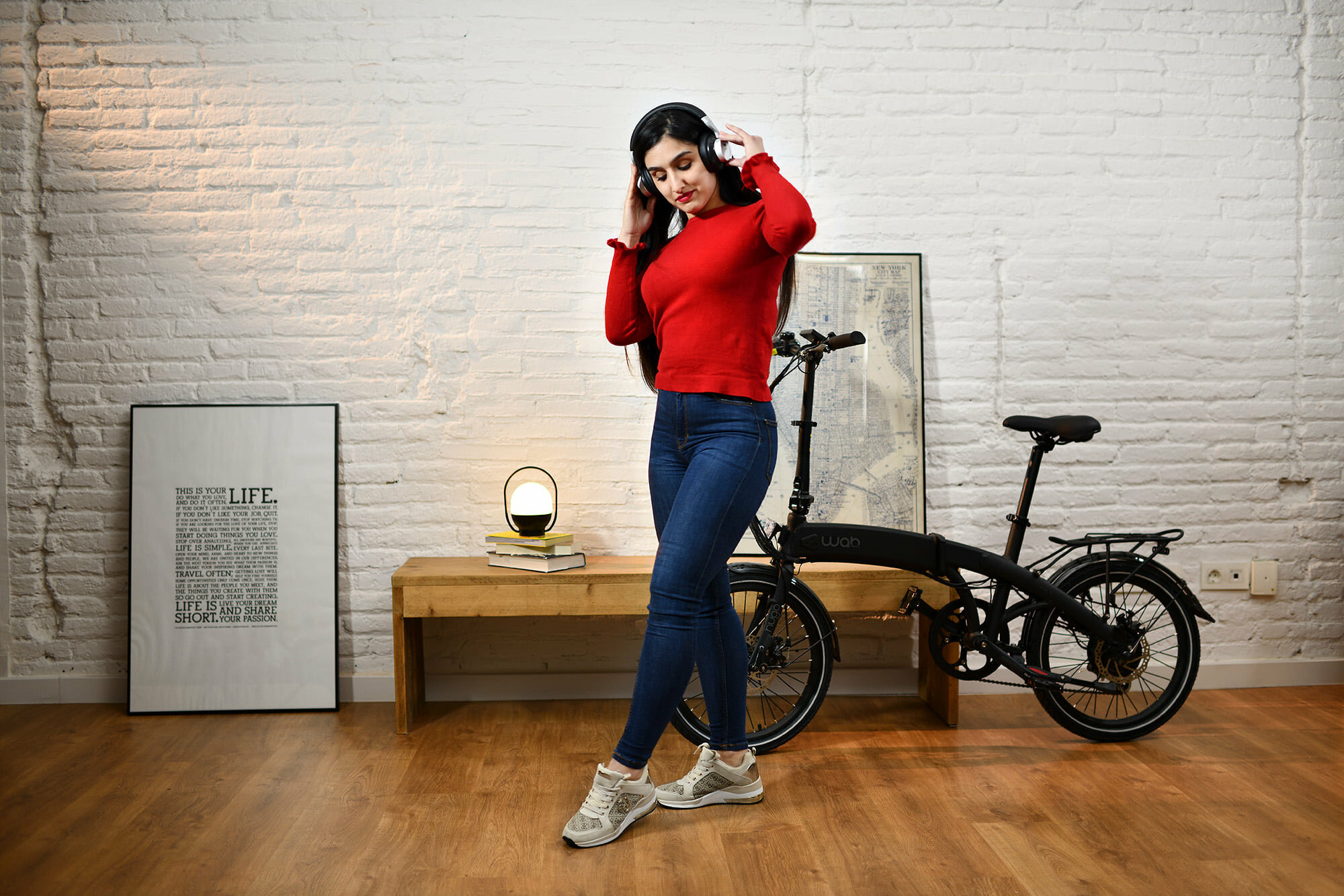 Una donna con cuffie vicino a una bici, con una panca in legno e un quadro lì vicino.