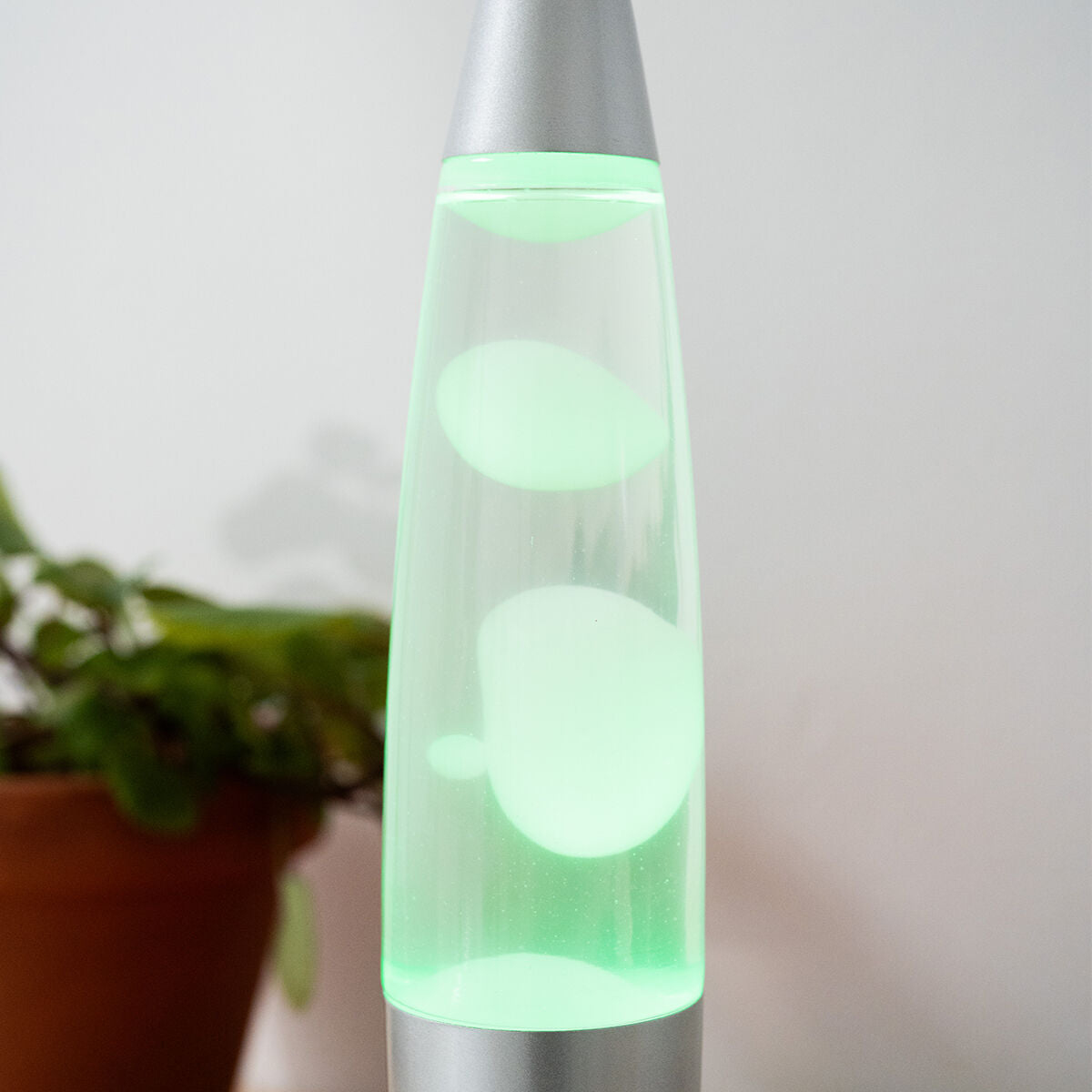 Una lámpara de lava verde con burbujas blancas dentro, contra una pared blanca con una planta.