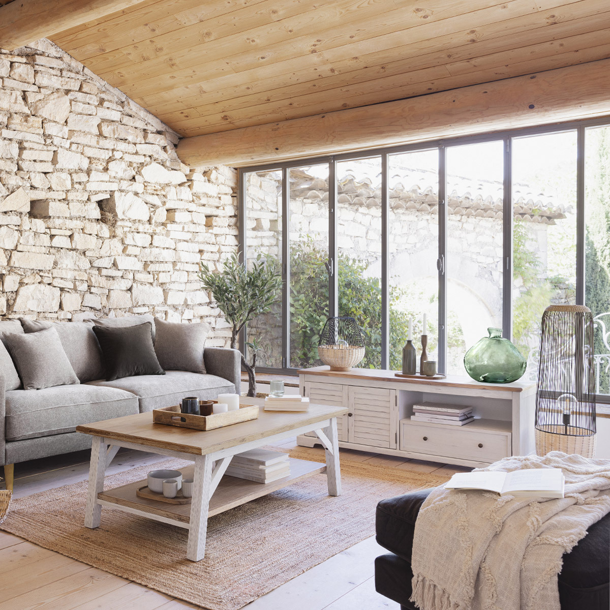 Salón con pared de piedra, techo de madera, grandes ventanales, sofá gris, mesa de centro y alfombra.