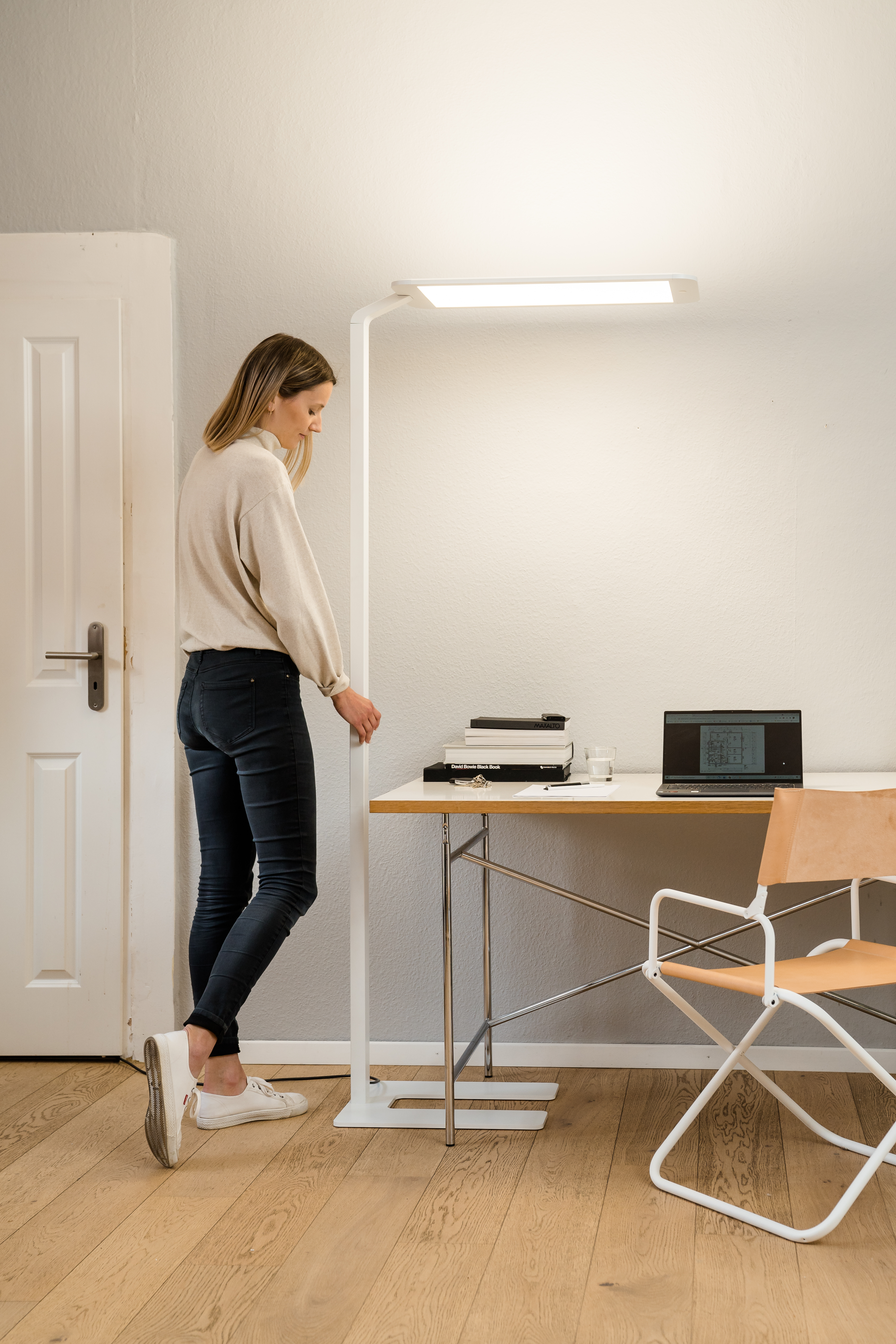 Frau neben einem Schreibtisch mit weißer Lampe. Es gibt Bücher und einen Laptop.