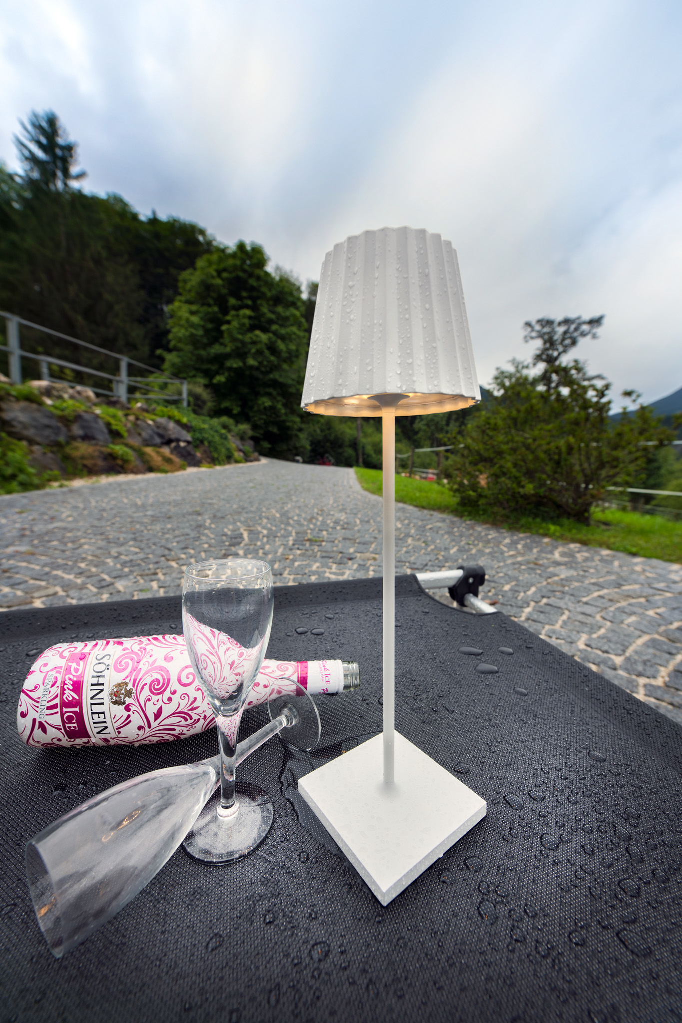 Una lampada bianca su un tavolo con una bottiglia di vino e bicchieri, scena all'aperto.