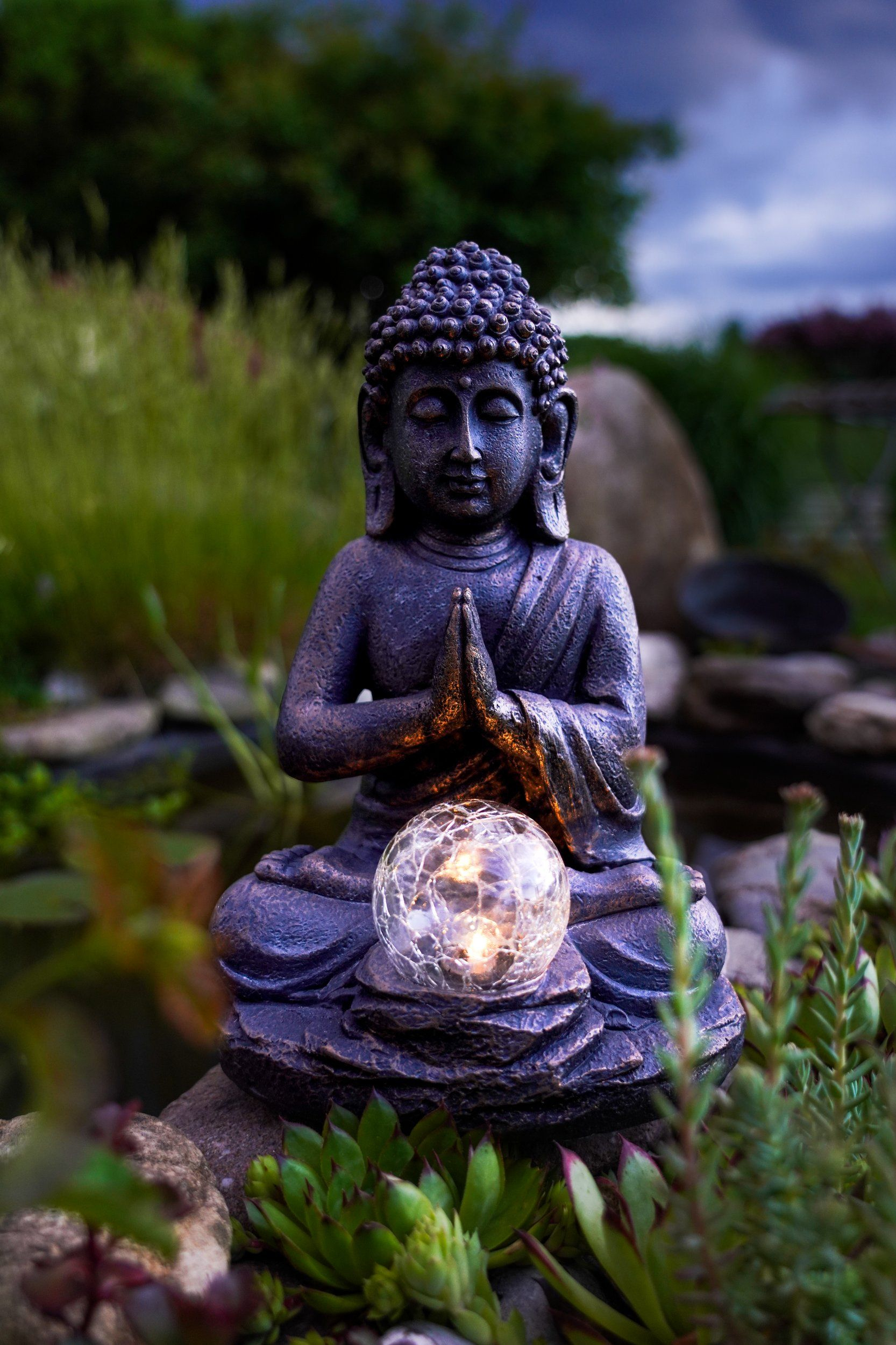 Buddha-Statue mit leuchtender Kugel. Hände im Gebet. In einem Garten mit Pflanzen aufgestellt.