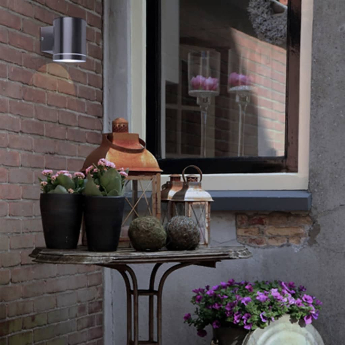 Scena all'aperto con un muro, una lampada, un tavolo, piante e fiori. Giorno, soleggiato.
