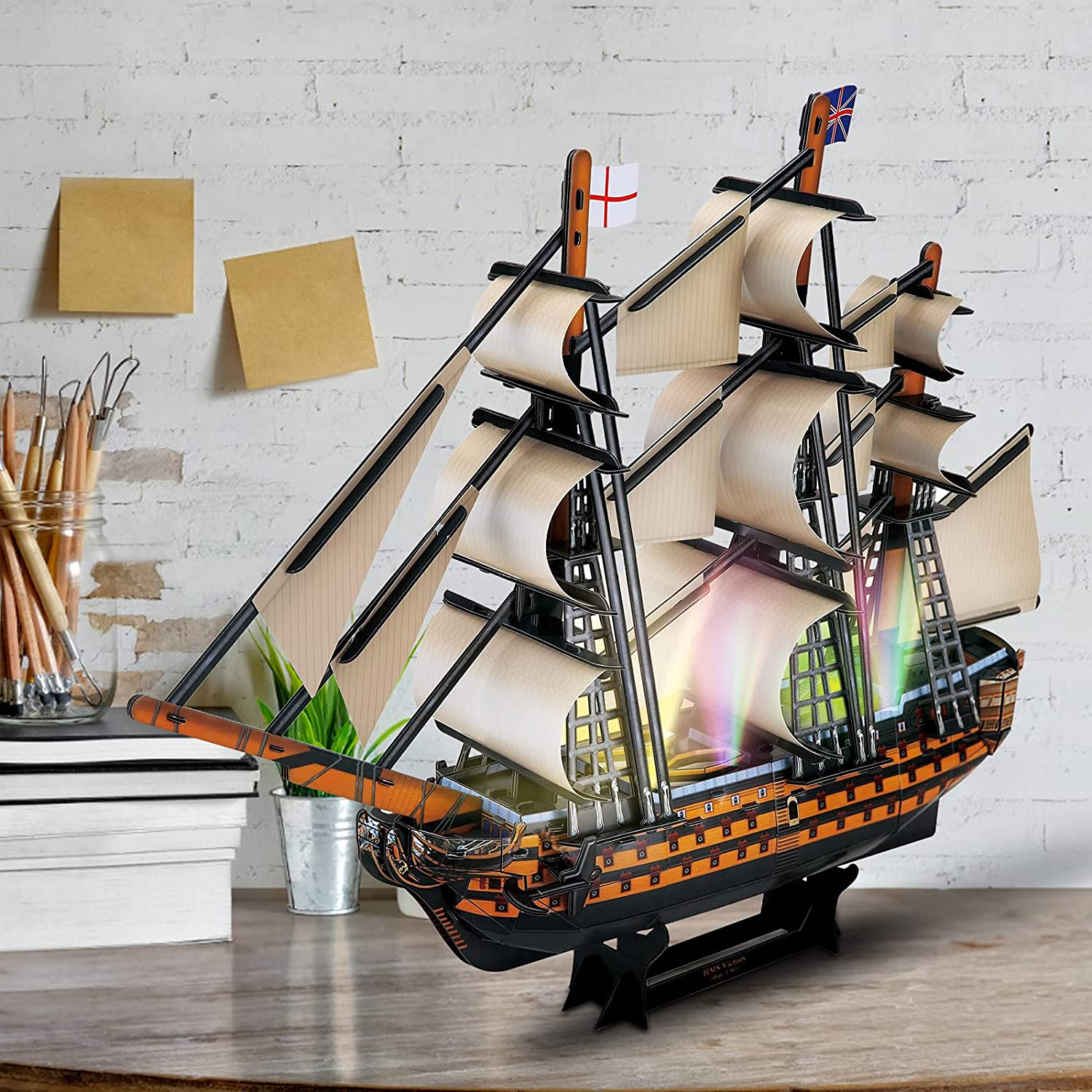 Ein Modellschiff auf einem Schreibtisch, mit Segeln und Flaggen. Im Hintergrund eine Backsteinmauer.