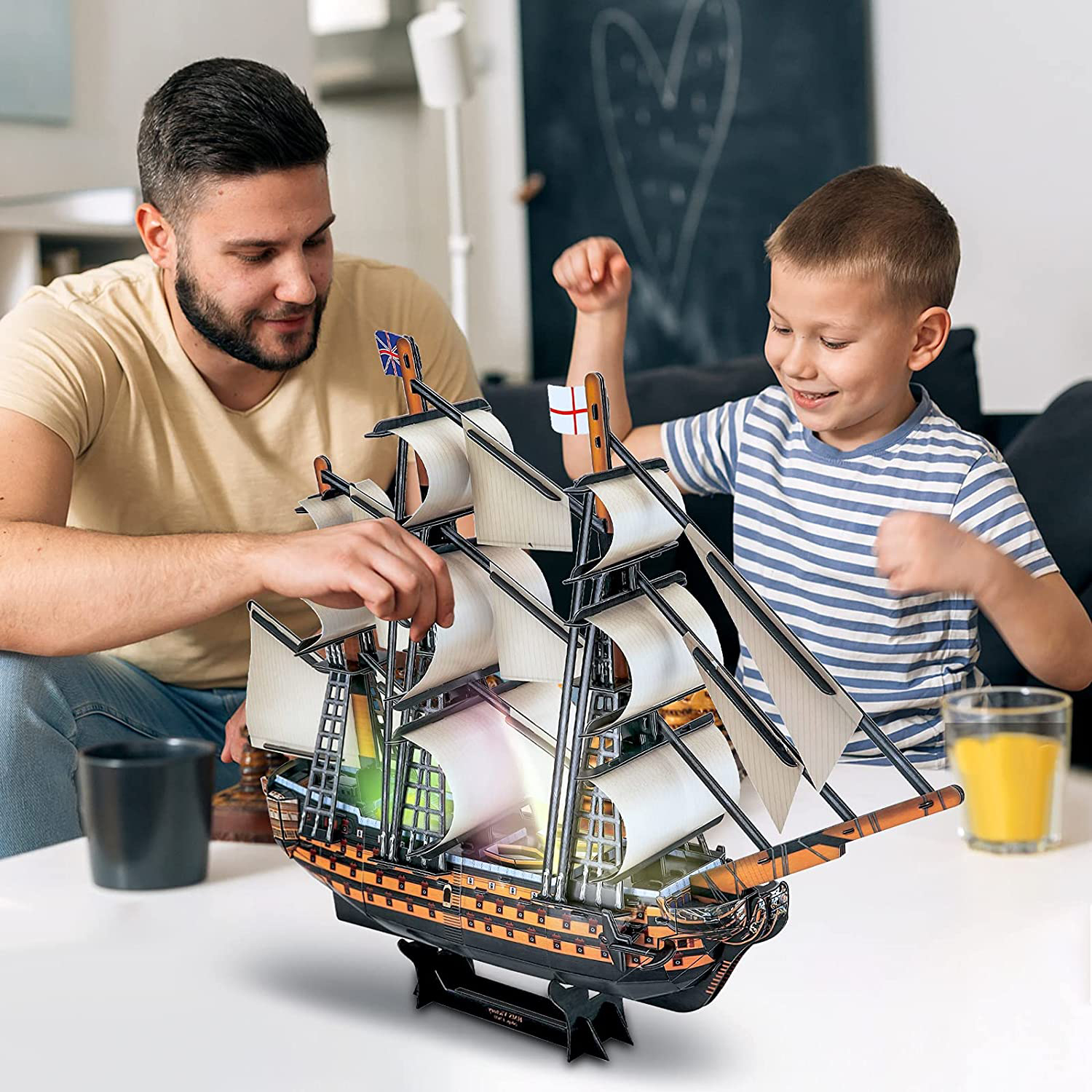 Vater und Sohn bauen ein Spielzeugschiff zusammen. Das Schiff steht auf einem weißen Tisch. Ein Glas Saft steht auf dem Tisch.
