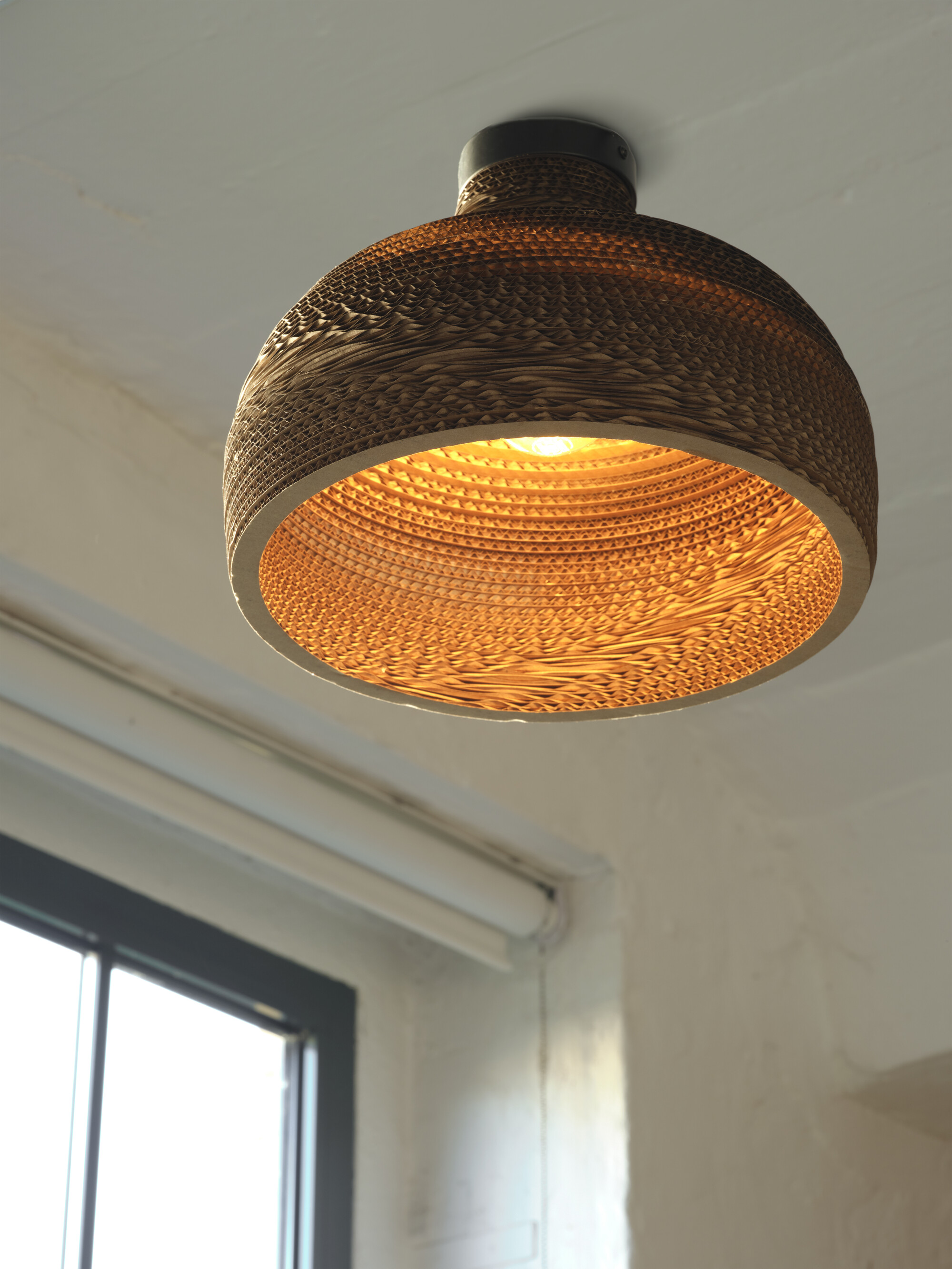 Hängende Papplampe mit Glühbirne. Weiße Decke und Wand sichtbar.