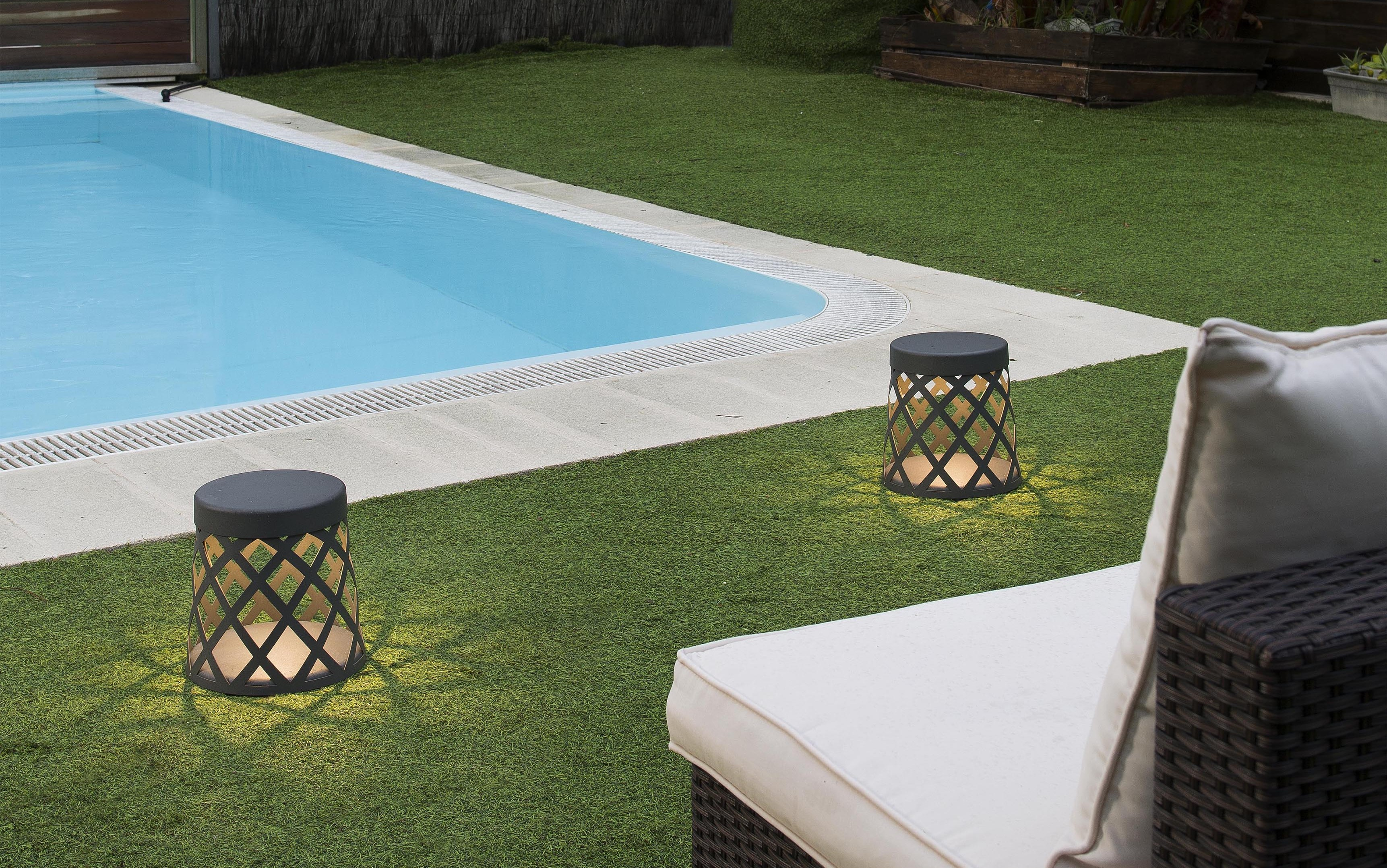Scena a bordo piscina: piscina, erba verde, due lanterne e una sedia a sdraio con cuscino bianco.