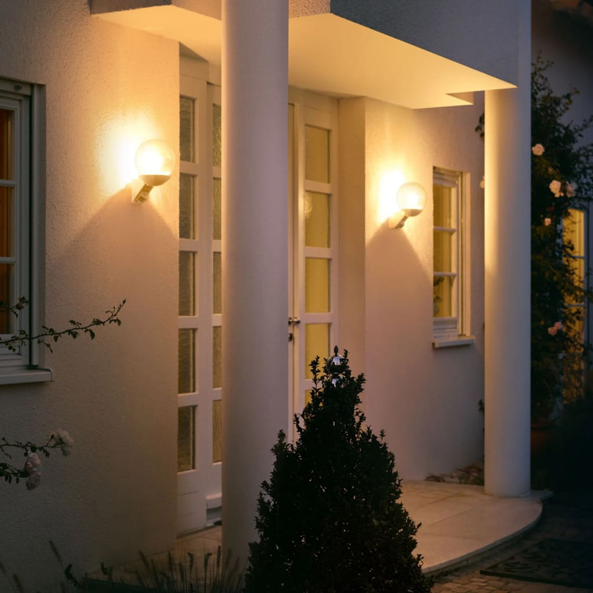 Hausfassade mit zwei Wandleuchten, Tür und Säulen, mit Garten und Büschen.