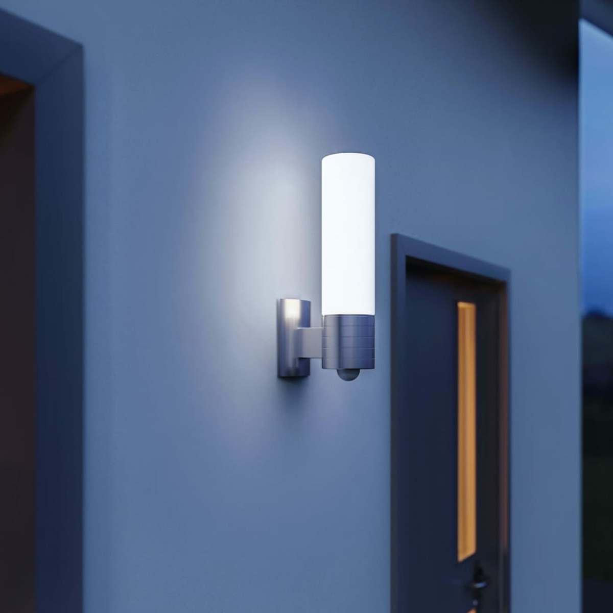 Una lampada cilindrica da esterno su un muro blu. La lampada è bianca e argento. Una porta sullo sfondo.