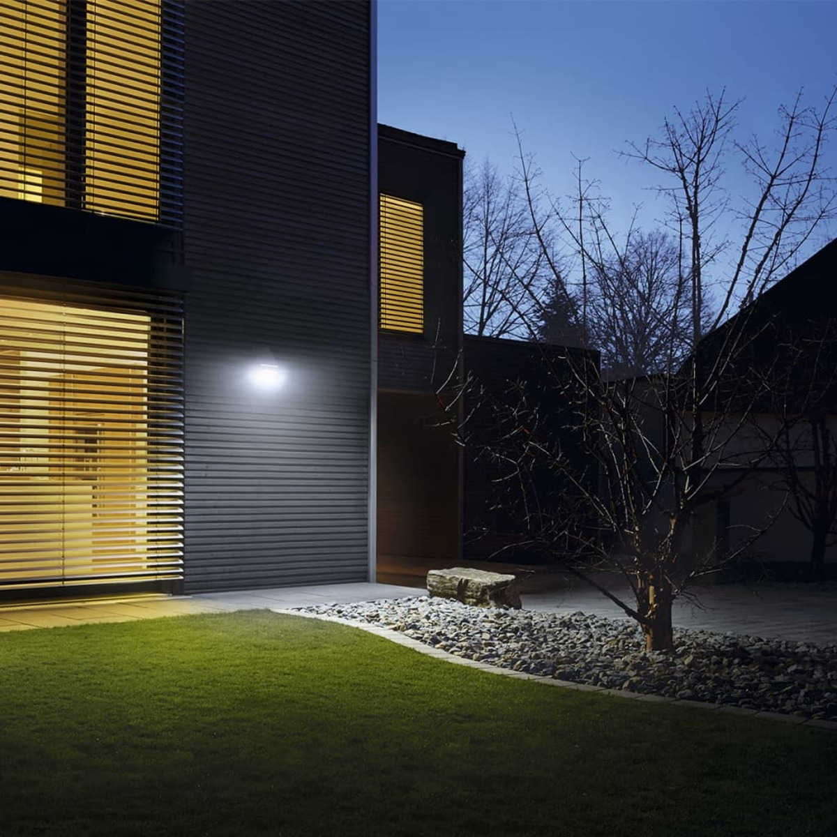 Hausfassade in der Dämmerung, beleuchtet von einer Wandleuchte. Grünes Gras, ein Baum und ein felsiger Bereich sichtbar.