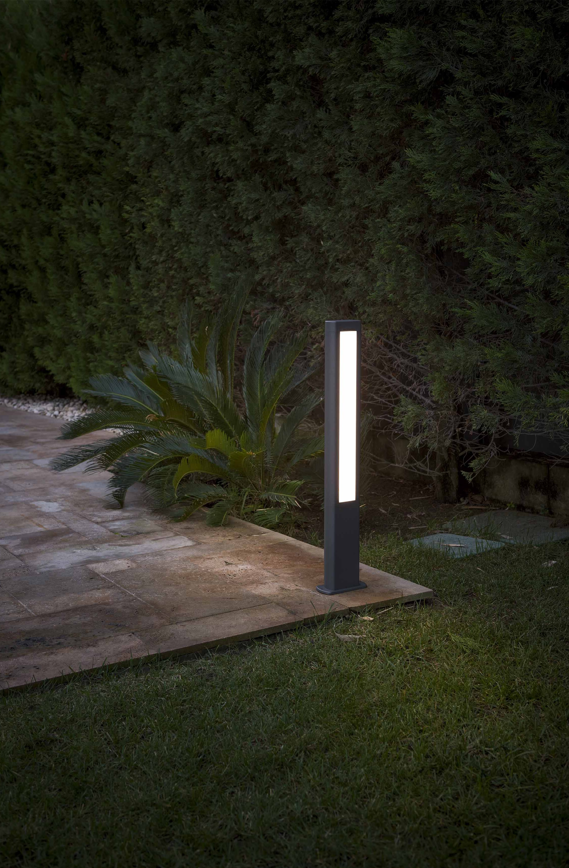 Una lampada da esterno rettangolare grigio scuro è su un sentiero di pietra, con erba e cespugli sullo sfondo.