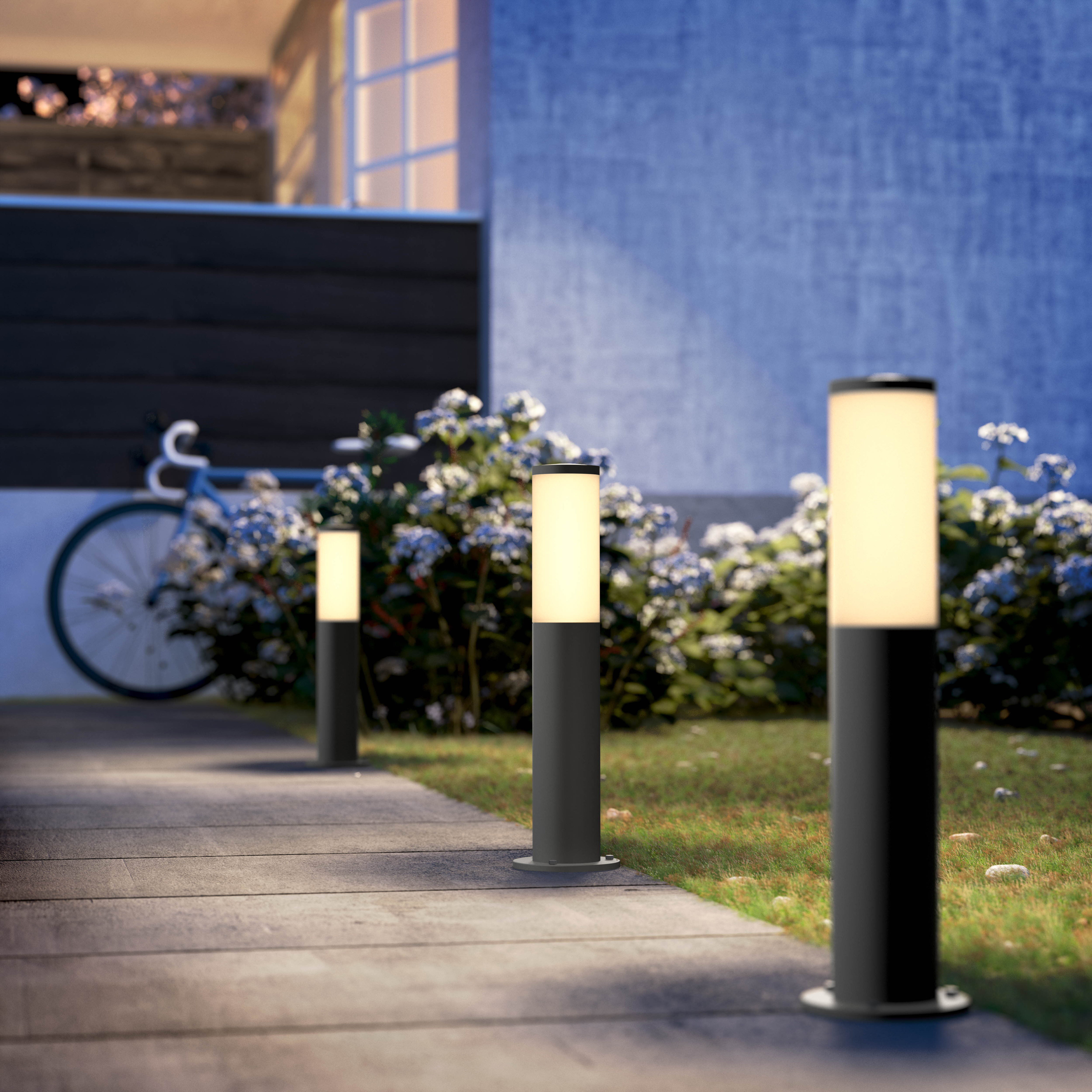 Tre luci cilindriche nere illuminano un vialetto di notte. Piante e una bicicletta sono sullo sfondo.