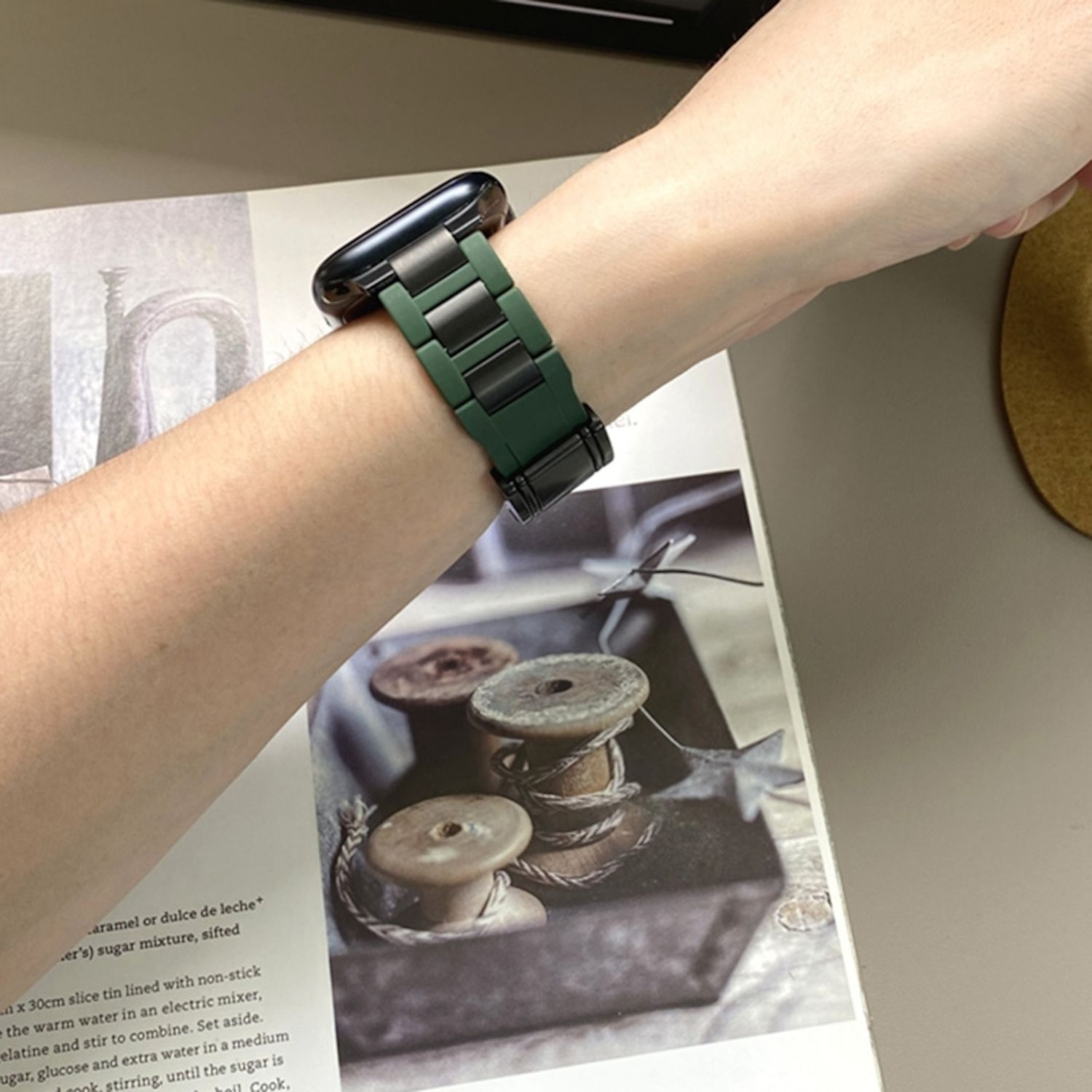 Handgelenk mit grünem Uhrenarmband auf einer Magazinseite mit Nähgarnrollen.