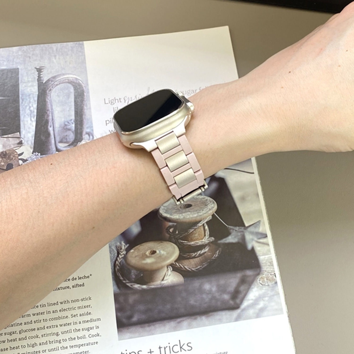 Handgelenk mit goldener Uhr und rosa Metallband, auf einem Magazin liegend.
