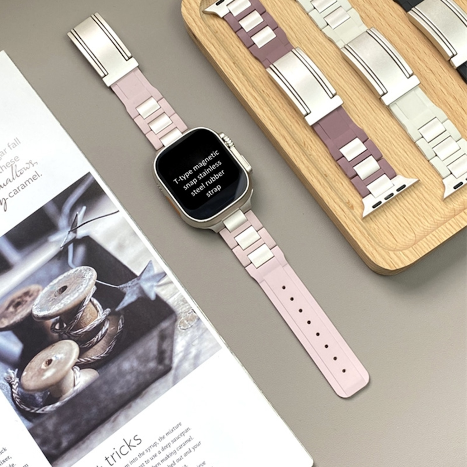 Eine Smartwatch mit pinkfarbenem Armband neben anderen Armbändern. Auf dem Tisch liegt eine Zeitschrift.
