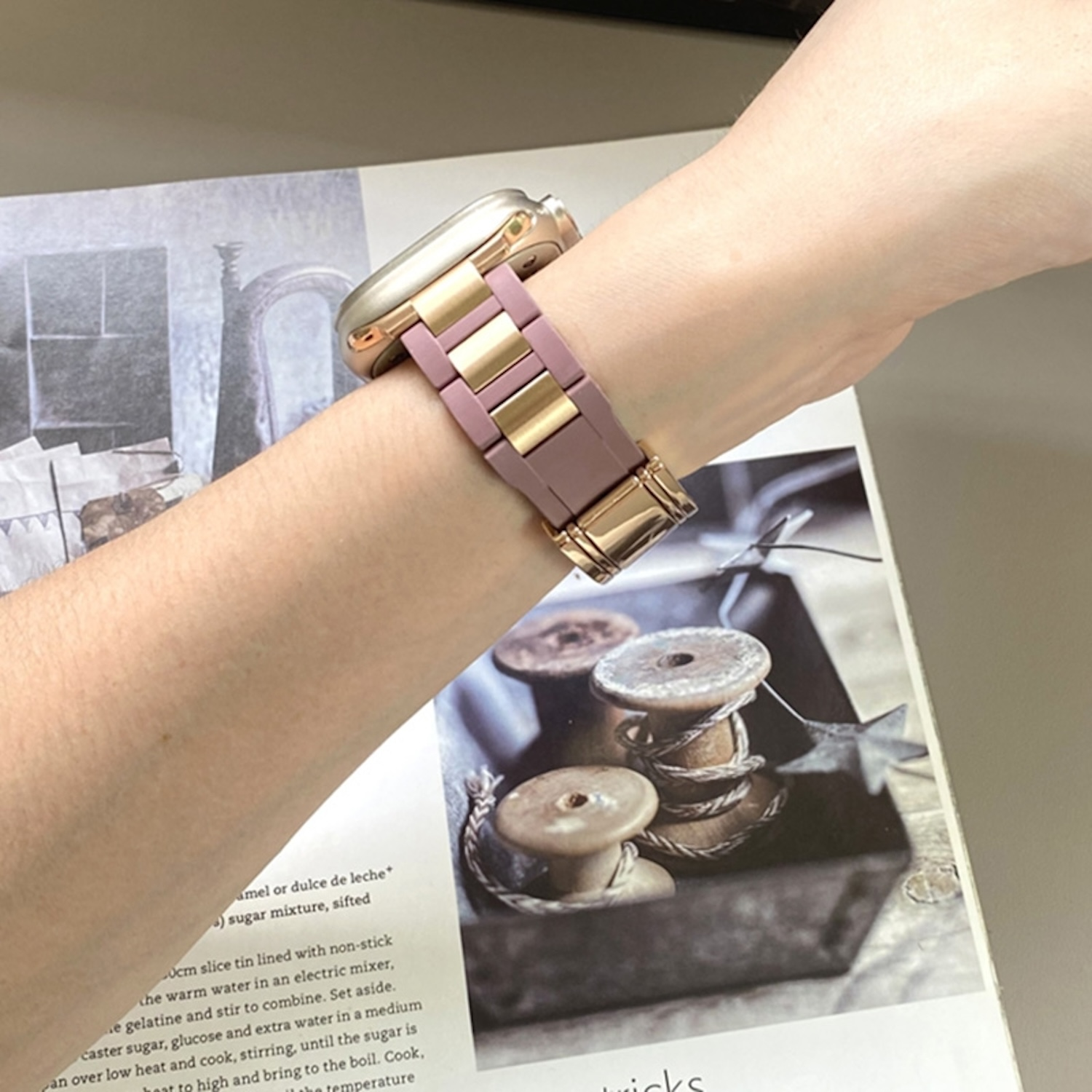 Das Handgelenk einer Person mit einem gold-lila Uhrenarmband auf einem Tisch mit einem Buch und Spulen.
