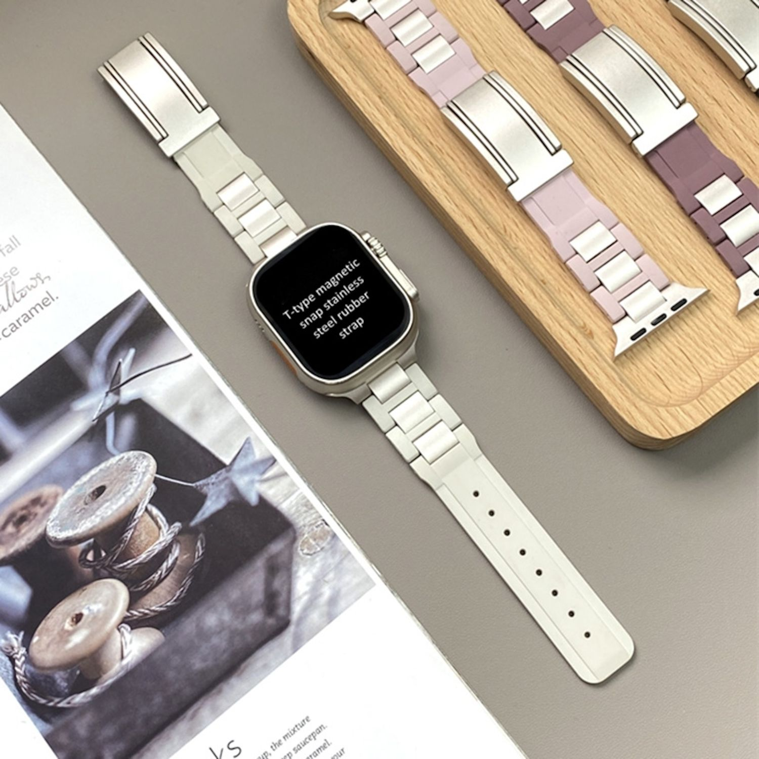 Weiße Apple Watch mit weißem und silbernem Armband auf hellgrauer Oberfläche neben einer Zeitschrift.