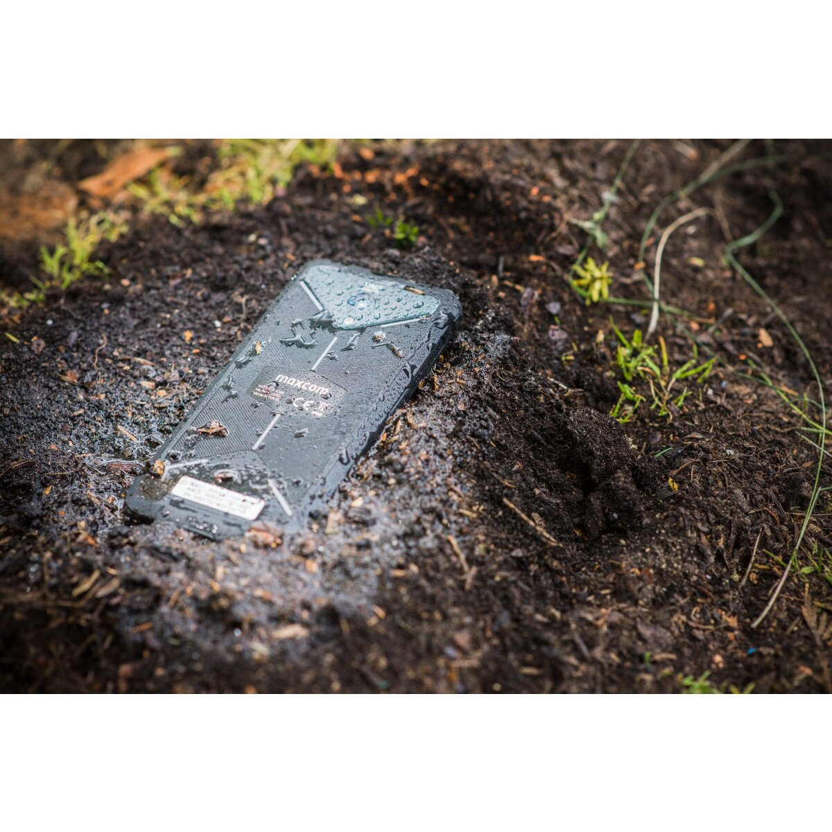 Robustes Telefon im schlammigen Boden, zeigt die Rückseite mit Markennamen.