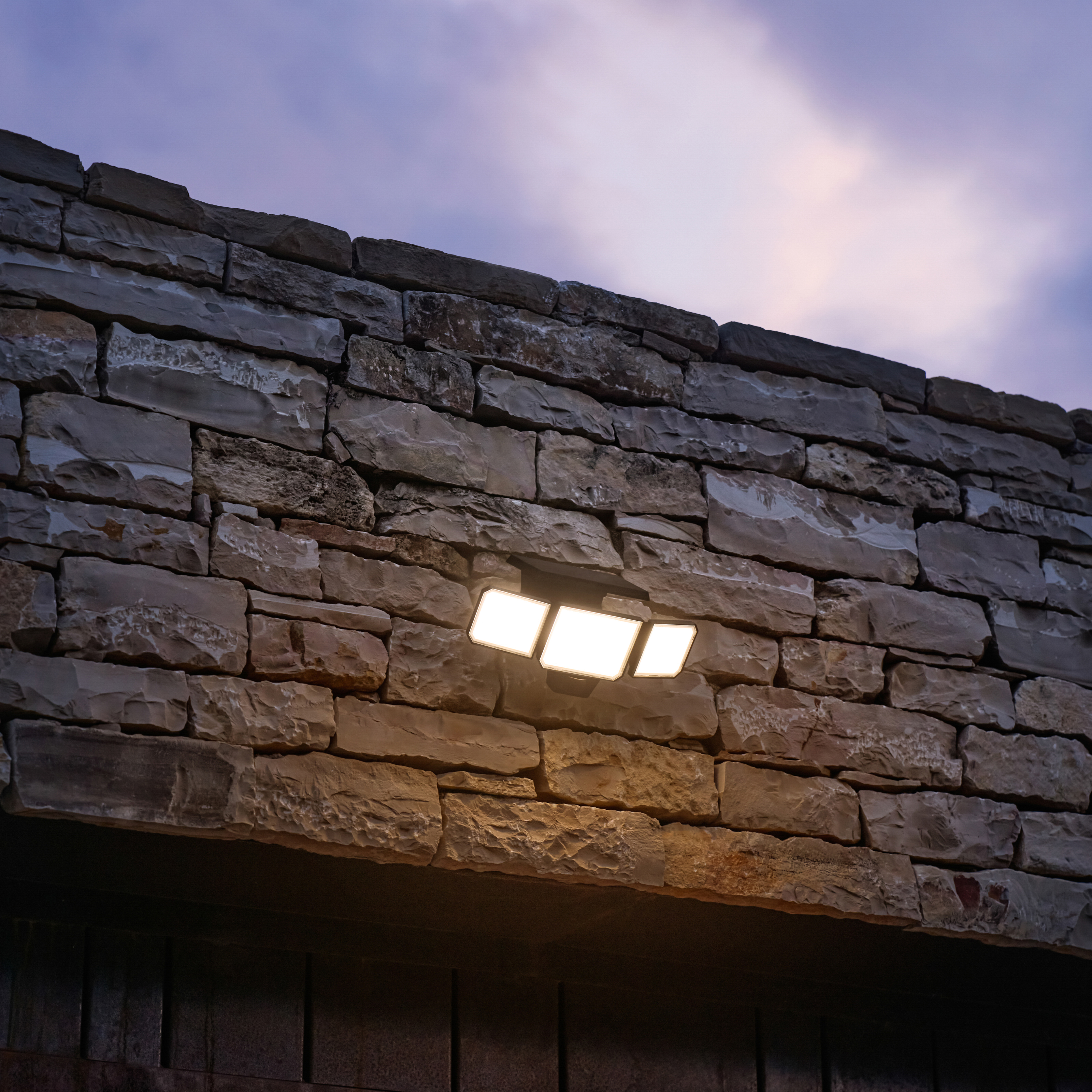 Lampada LED a tre luci su muro in pietra, illuminata, contro cielo nuvoloso.