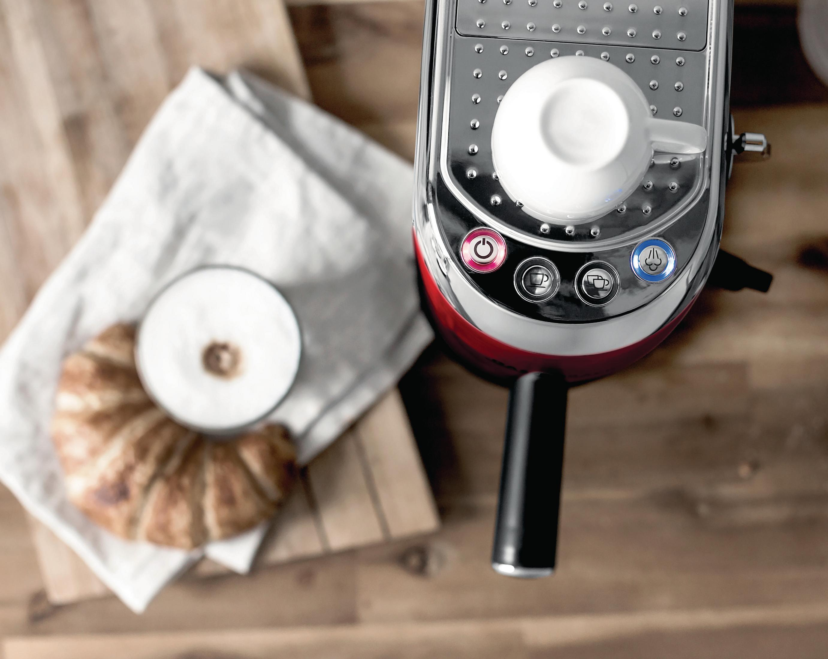 Ekspres do kawy z filiżanką na górze, rogalikiem i filiżanką piany, na drewnianym stole.