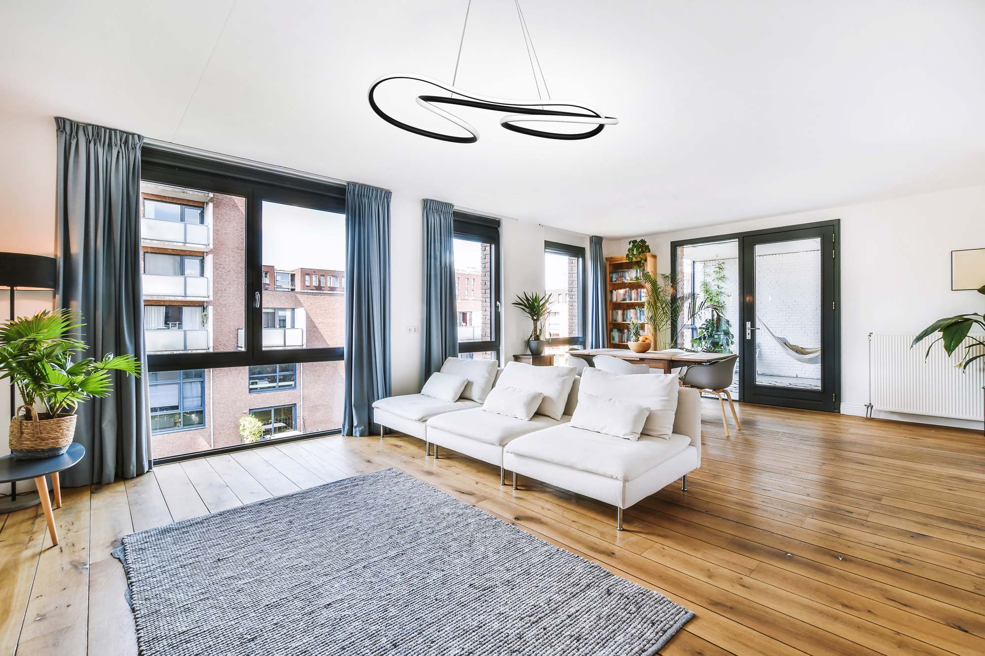 Escena de salón moderno con sofá blanco, alfombra y grandes ventanas con vistas a edificios.