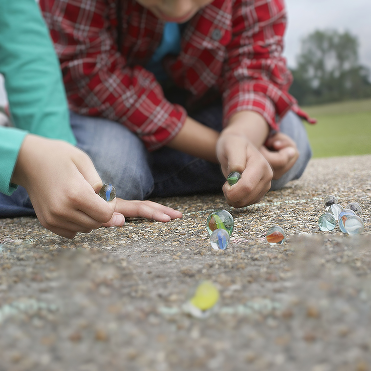 Bambini che giocano a biglie su cemento, erba verde sullo sfondo.