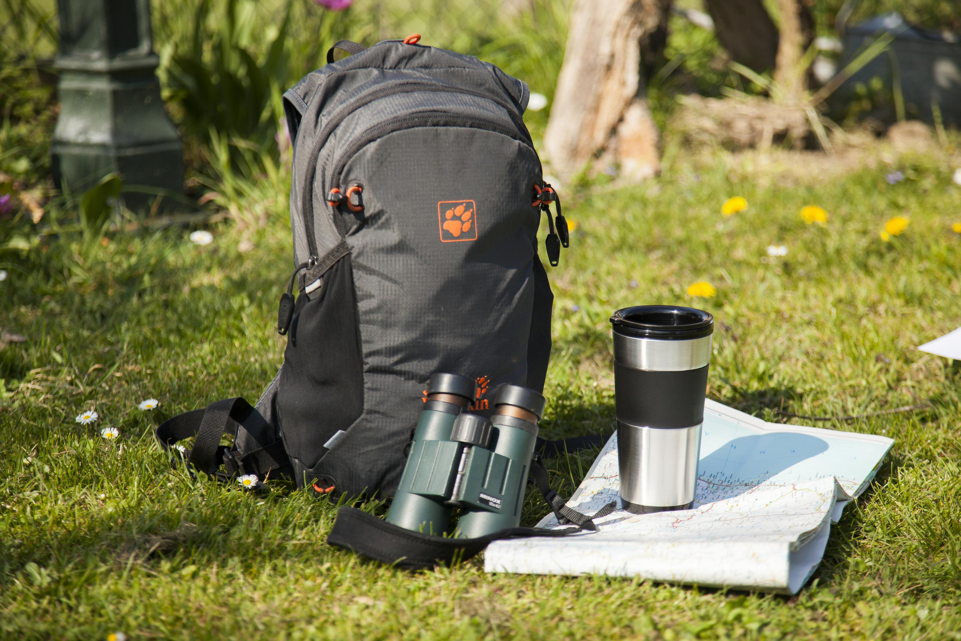 Plecak, lornetka, mapa i termos na trawie, w świetle słonecznym.