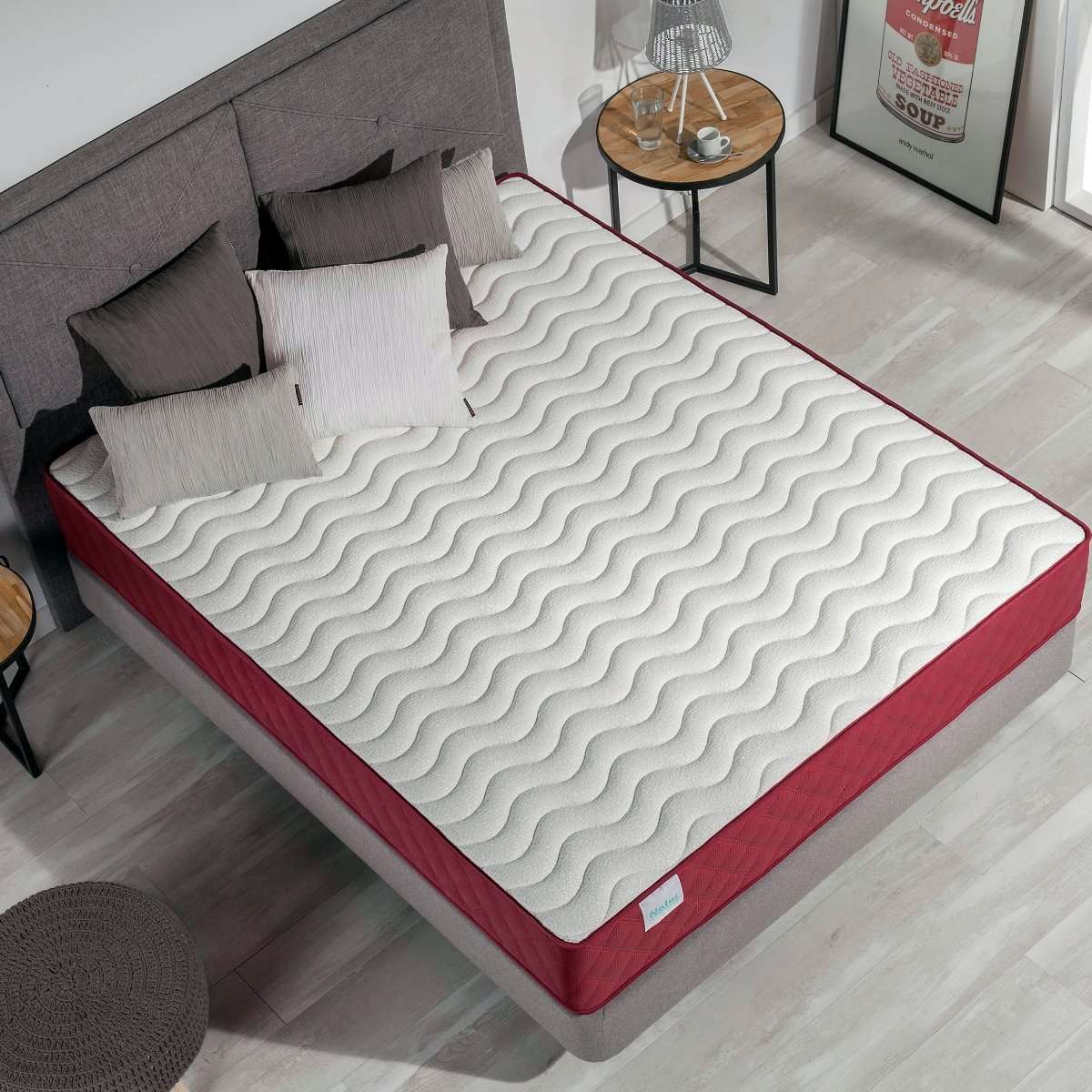 Un colchón sobre un marco de cama con almohadas y una mesa auxiliar con una lámpara.