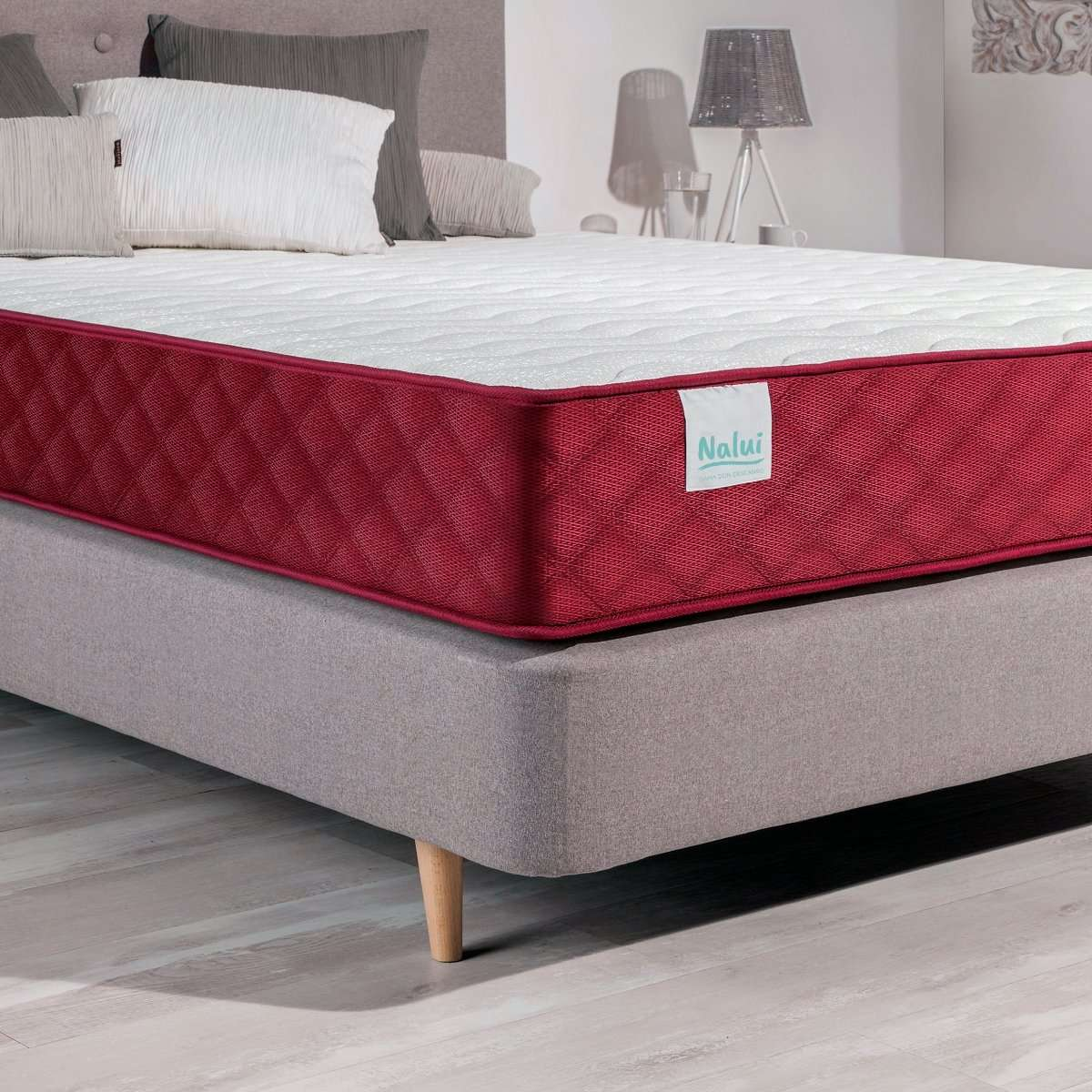 Cama con colchón blanco, borde rojo y base gris, con almohadas y lámpara en el fondo.