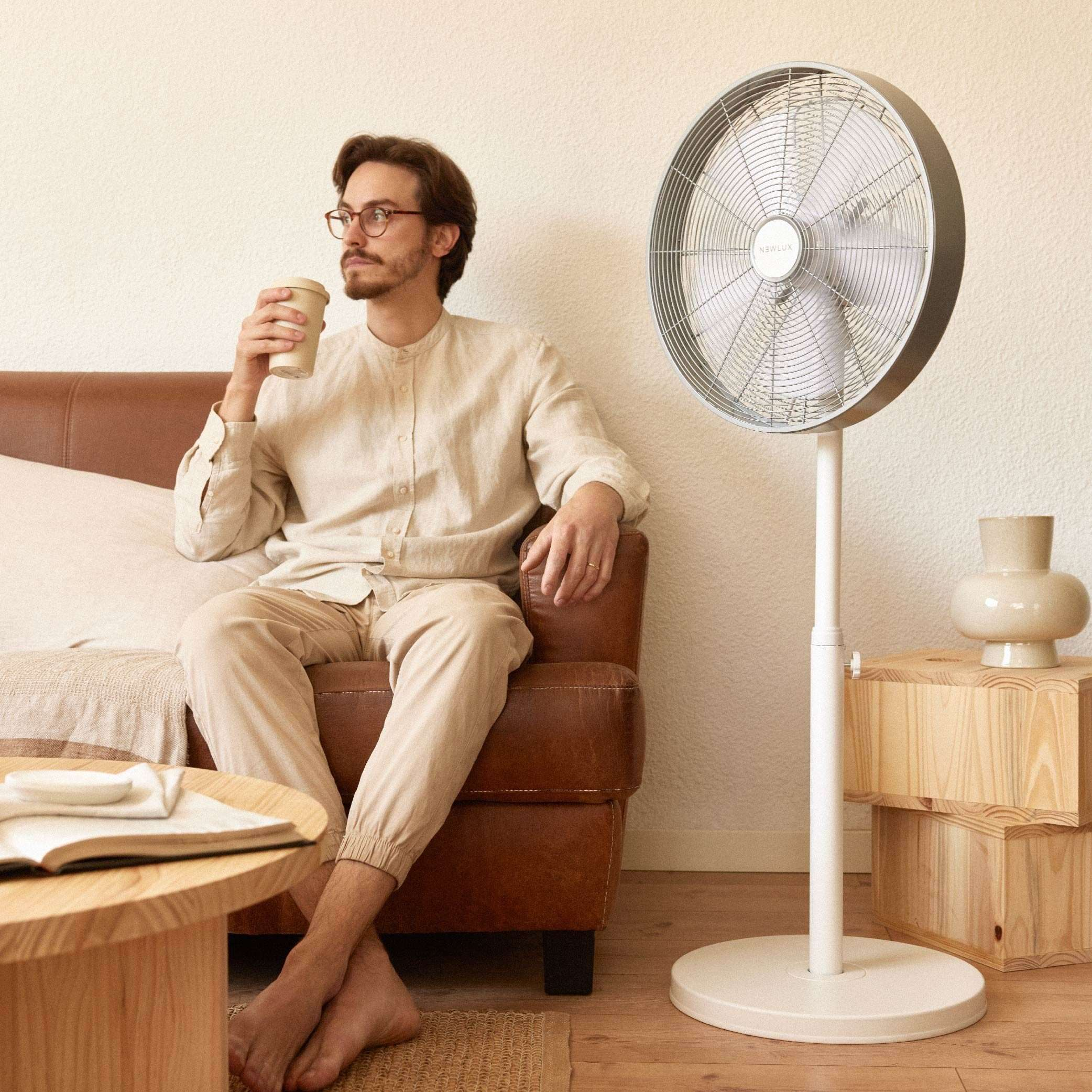 Un hombre con una taza de café sentado en un sofá junto a un ventilador de pie.