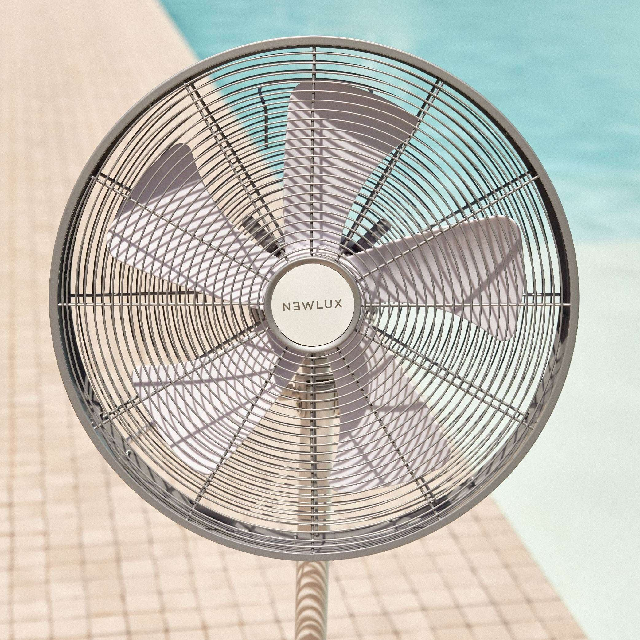 Ventilador plateado con jaula redonda frente a una piscina azul. El ventilador está en un poste blanco.