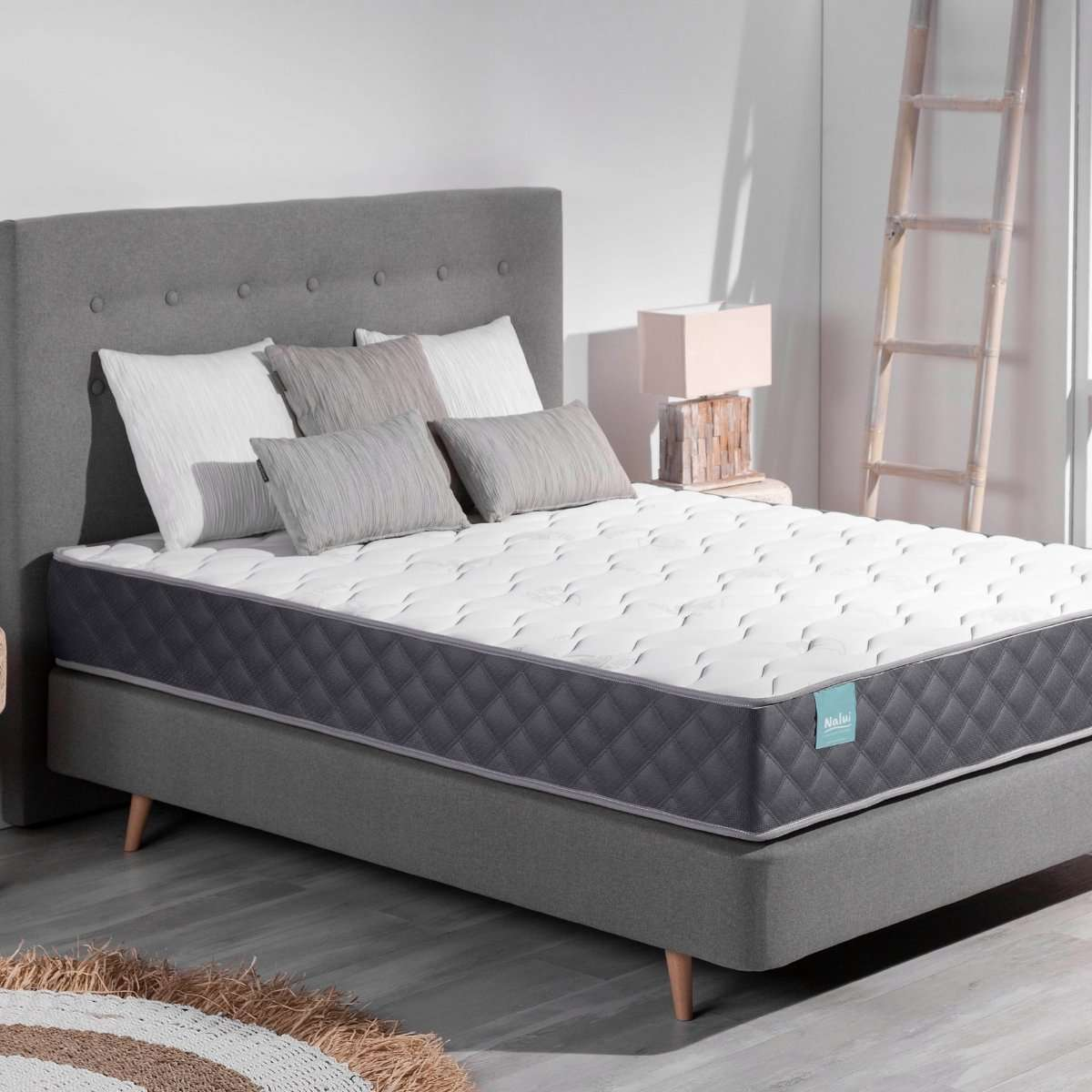 Un colchón blanco y gris sobre un somier gris con almohadas, una lámpara y una escalera.