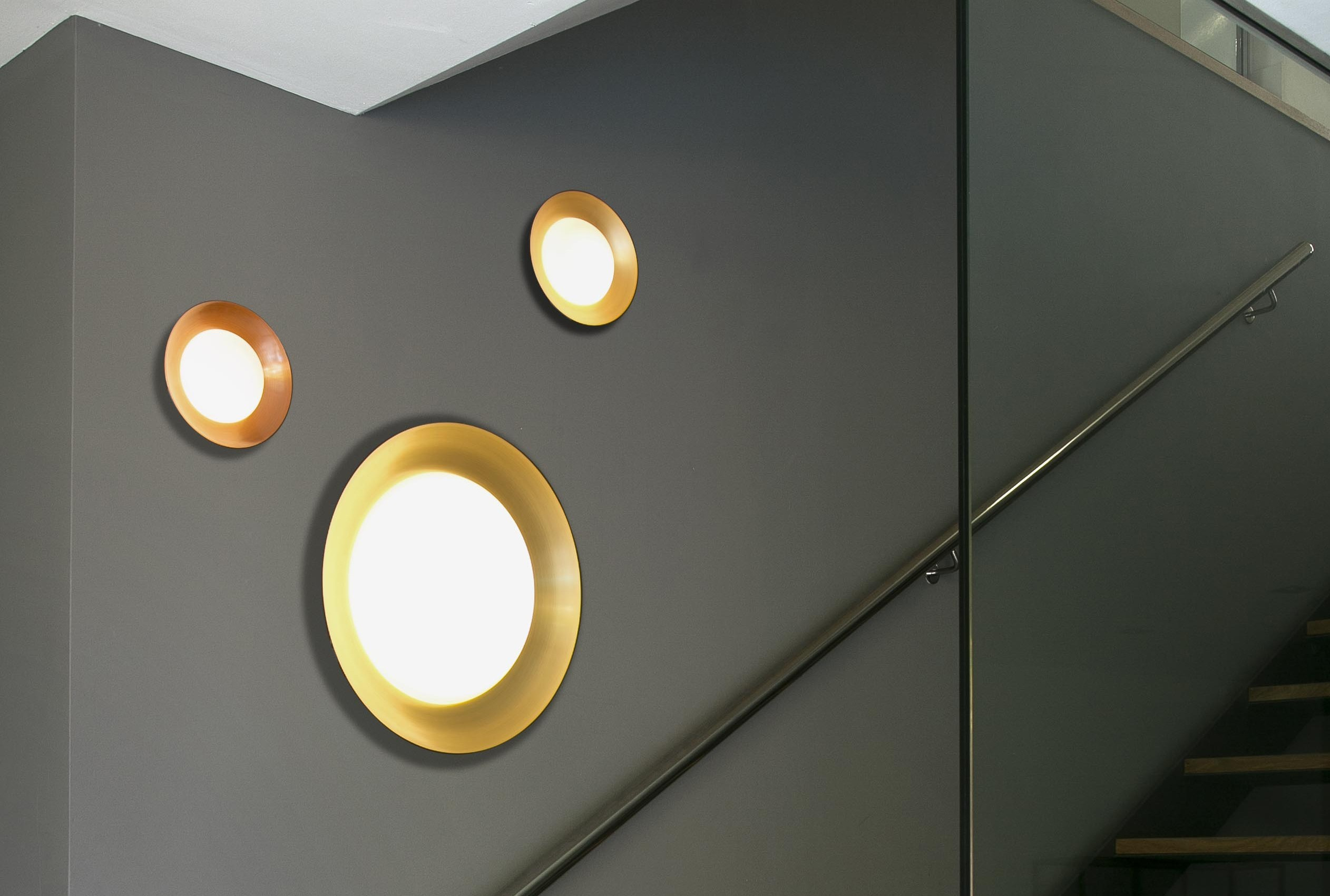 Tre applique con cerchi dorati, su un muro grigio scuro, con una scala sullo sfondo.