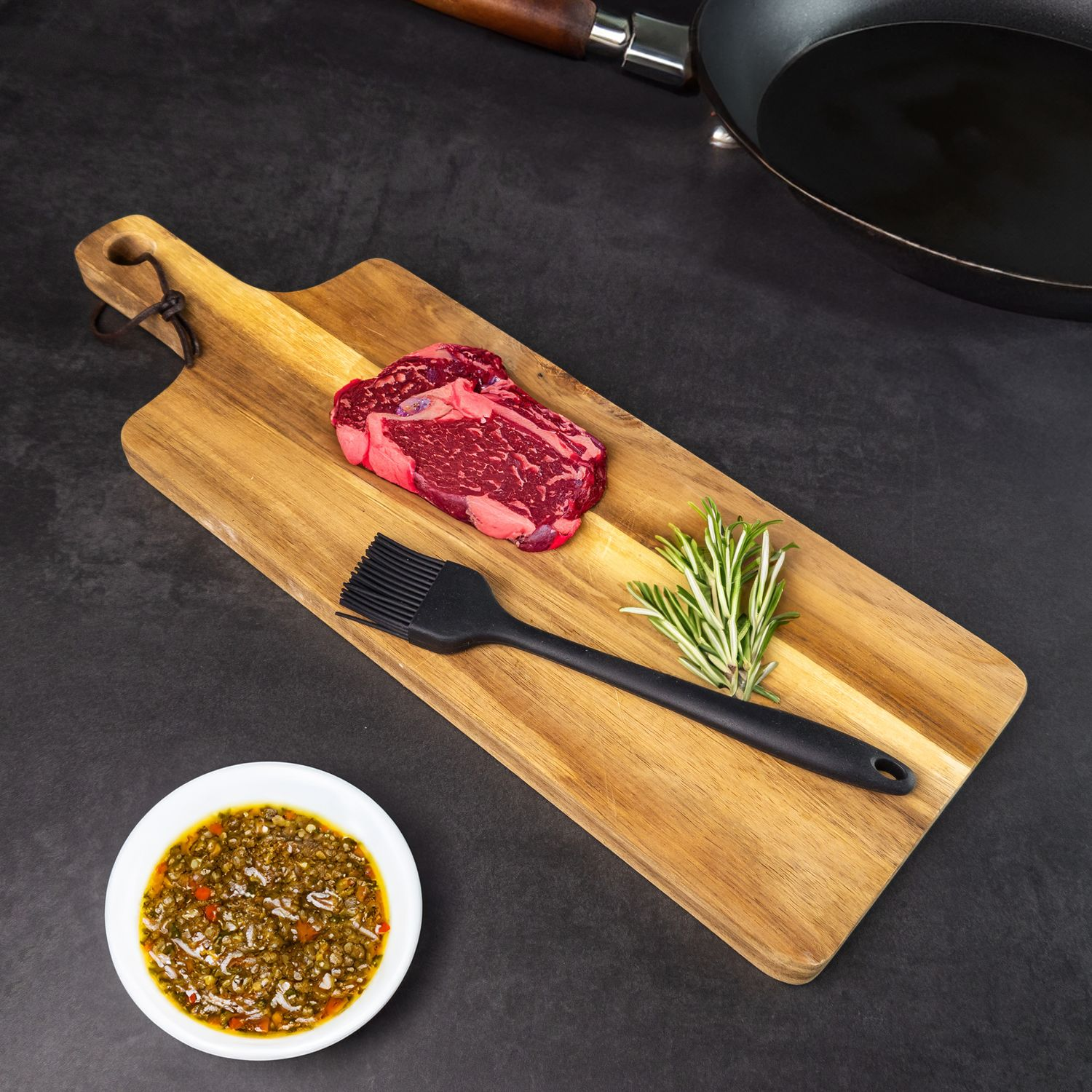 Holzschneidebrett mit Steak, Pinsel, Rosmarin und Soße auf schwarzer Oberfläche.