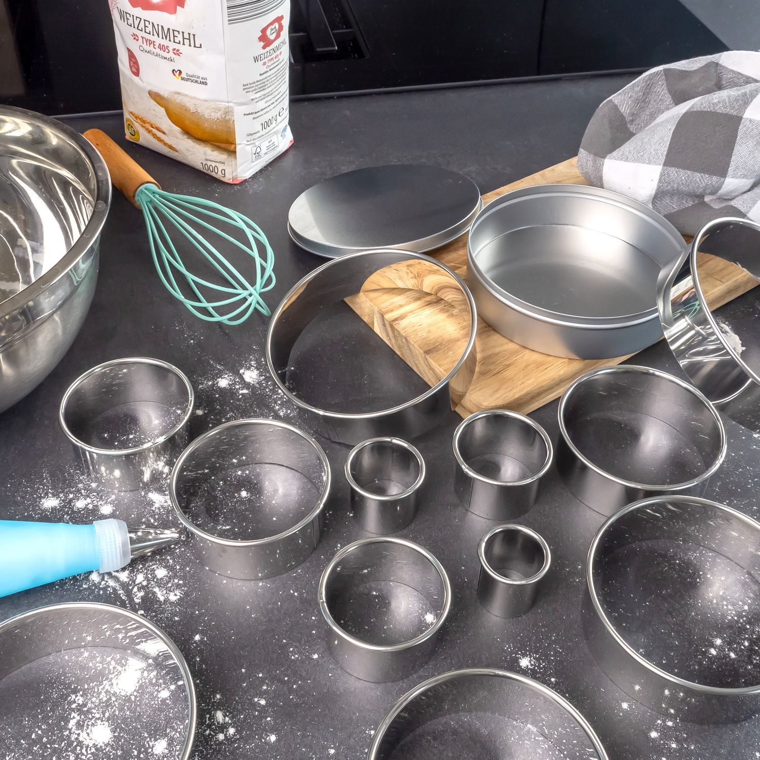Backszene: Backutensilien, Mehl und Backringe liegen auf einer schwarzen Oberfläche.