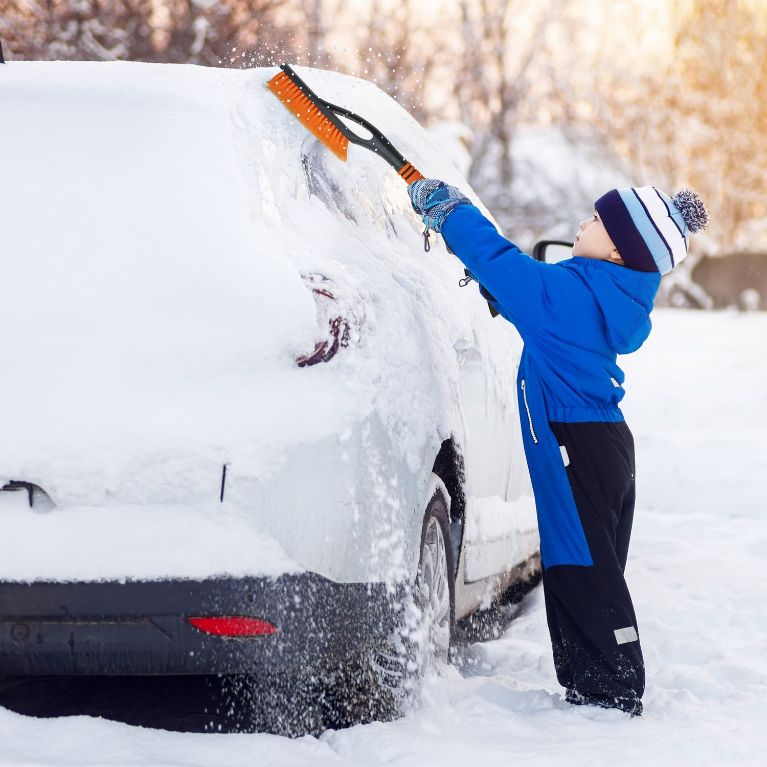 Ein Junge in einer blauen Jacke fegt Schnee mit einer Schneebürste von einem weißen Auto.