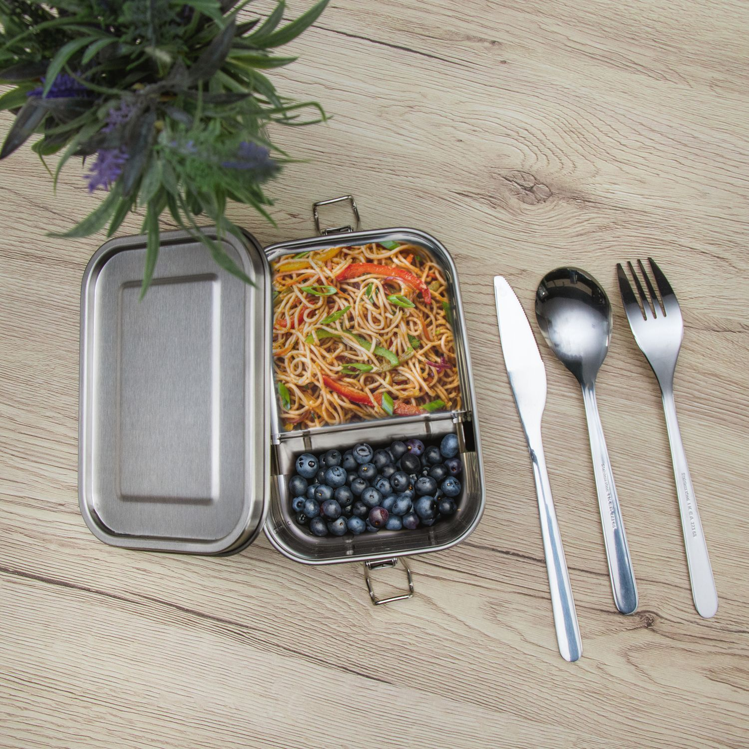 Edelstahl-Lunchbox mit Nudeln und Blaubeeren, mit Besteck auf einem Holztisch.