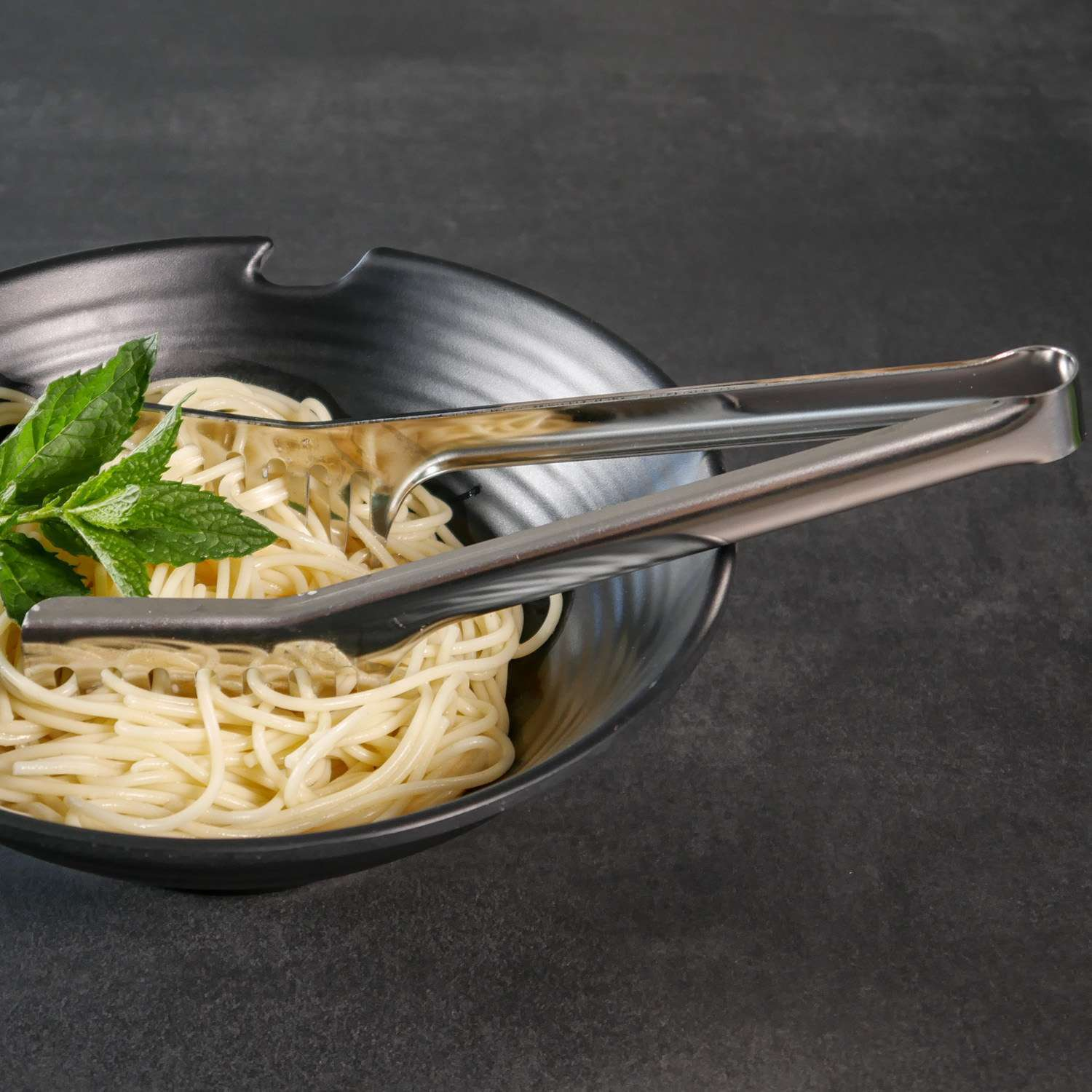 Spaghetti mit Minze in einer schwarzen Schüssel, silberne Zangen oben, dunkler Hintergrund.