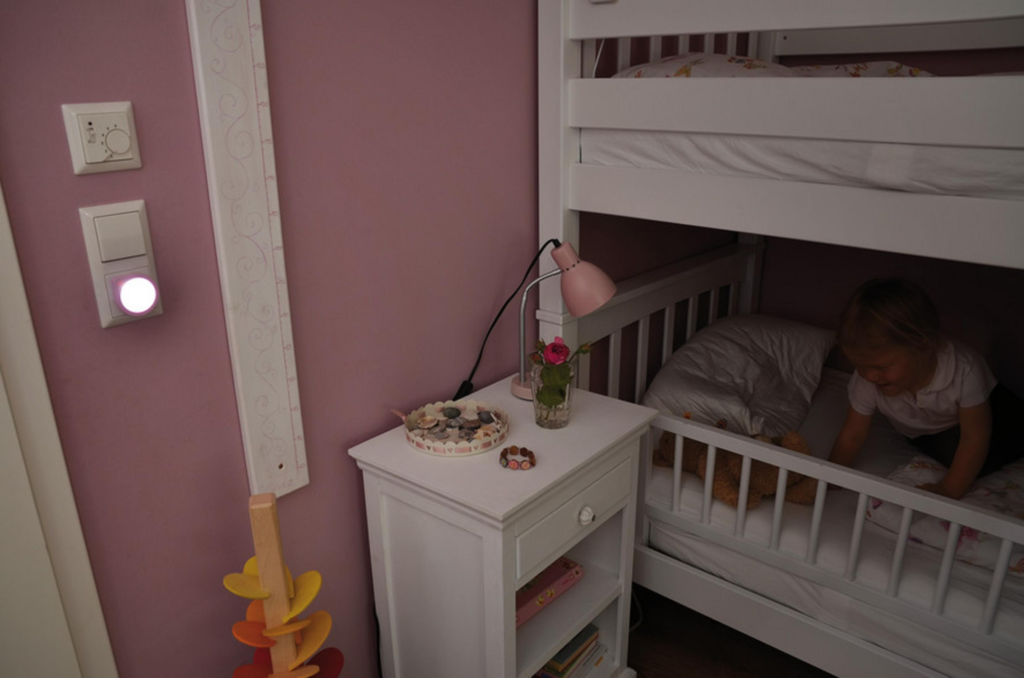 Pembe duvarlı ranza odasında bir çocuk. Bir lamba ve pencere bulunan bir komodin var.