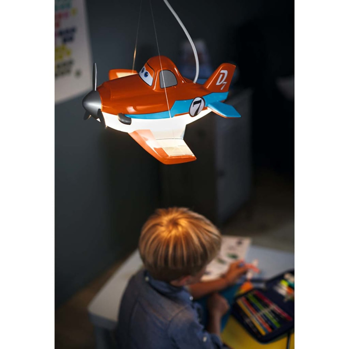 Un bambino disegna a un tavolo sotto una lampada a forma di aereo giocattolo. La lampada è arancione e blu.
