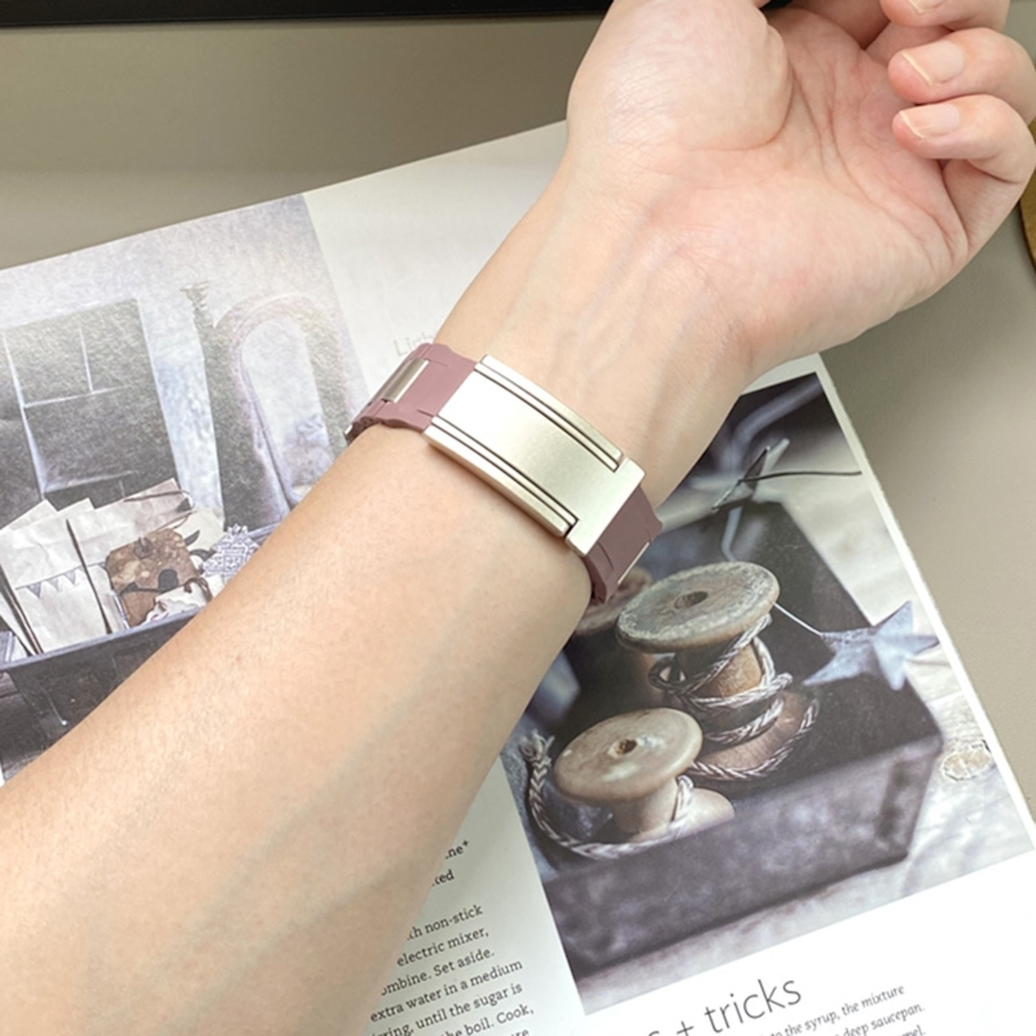 Das Handgelenk einer Person mit einem rosa-goldenen Uhrenarmband auf einem Magazin mit Holzspulen.