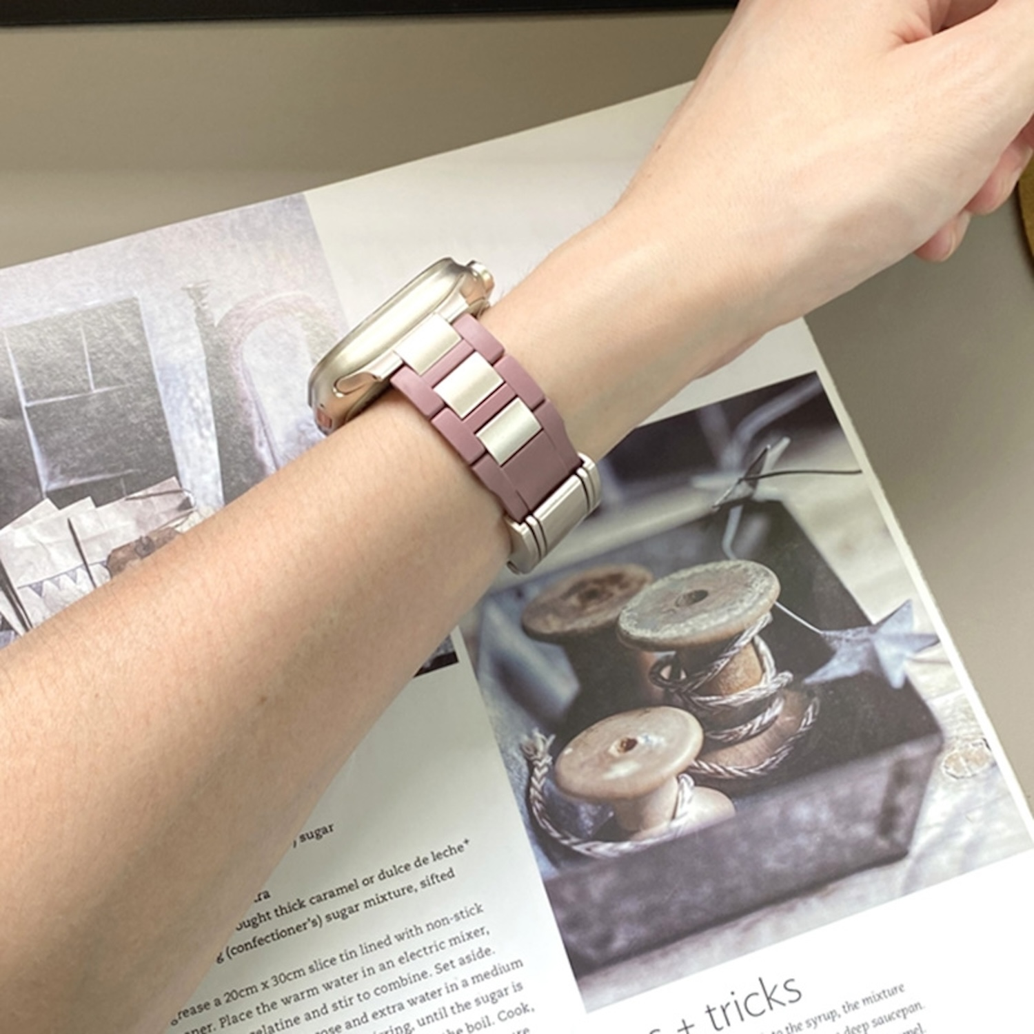 Handgelenk mit Uhr und pink-silbernem Armband. Zeitschrift auf dem Tisch.