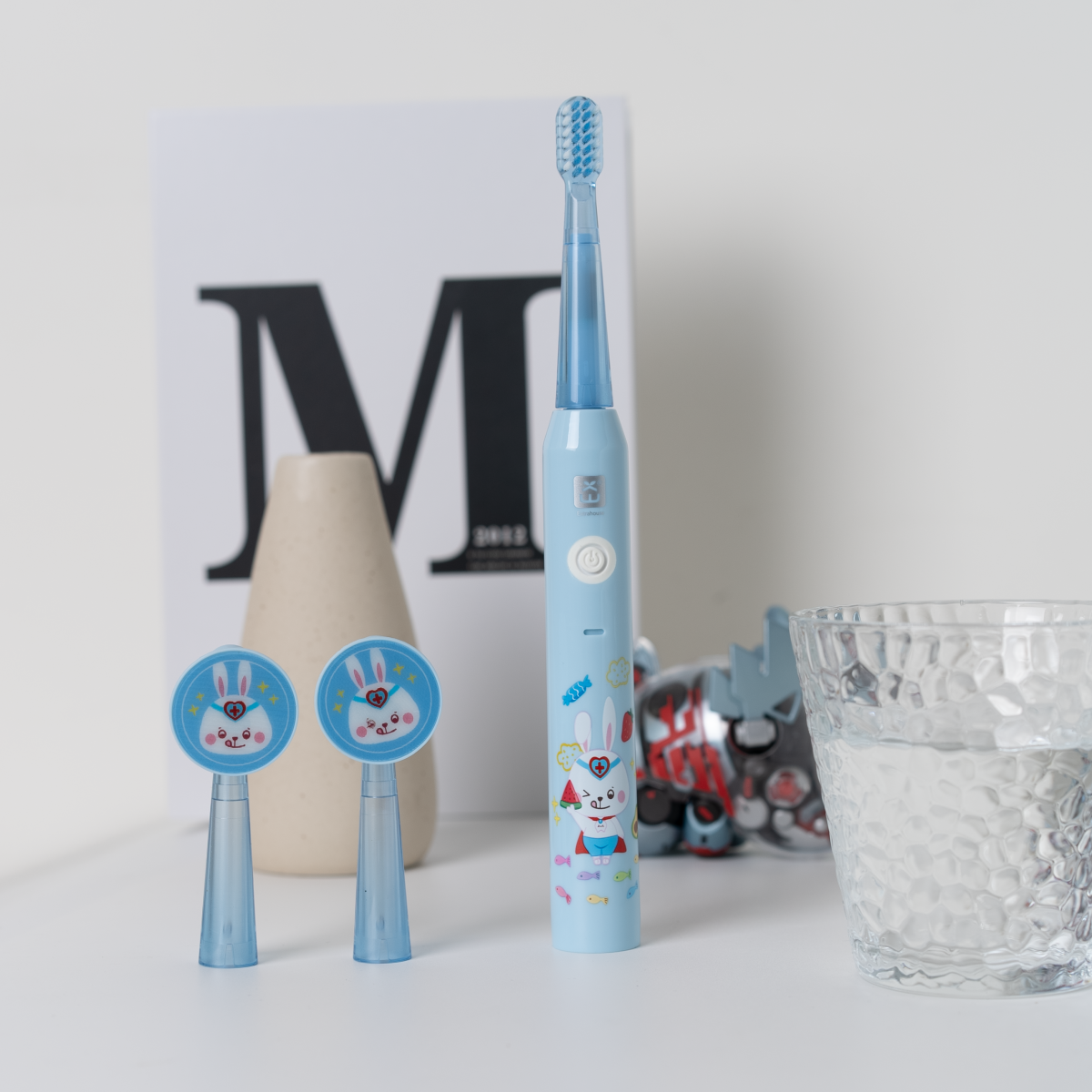 Un cepillo de dientes eléctrico azul y dos cabezales con diseños de conejos de dibujos animados, y un vaso.