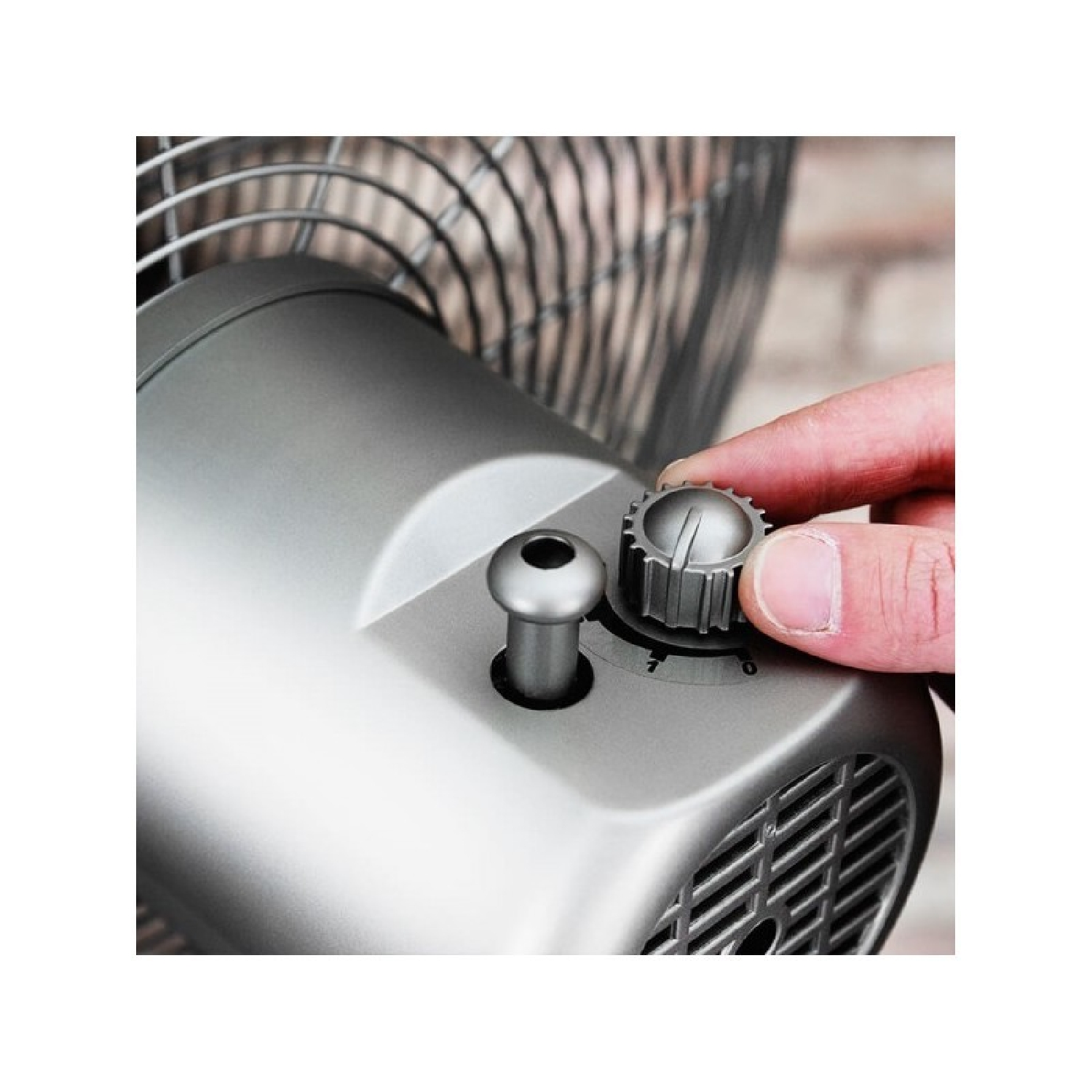 Una mano ajustando un dial de velocidad de ventilador plateado, con el ventilador al fondo.