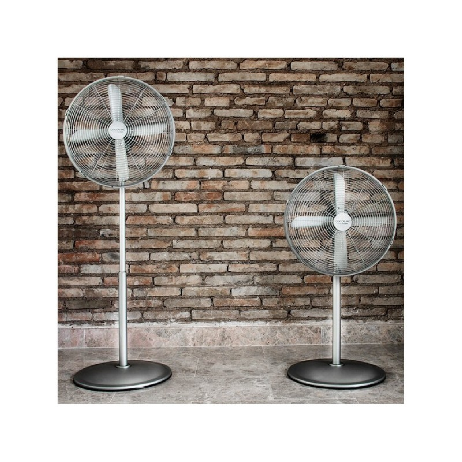 Dos ventiladores plateados sobre suelo gris, contra pared de ladrillos.