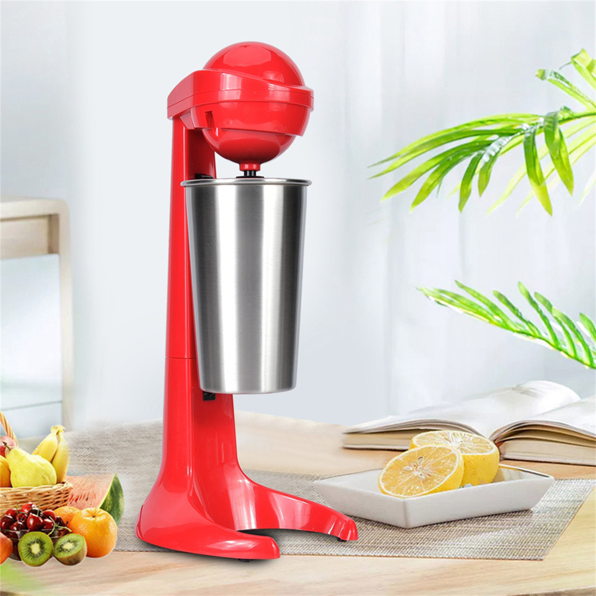 Roter Milchshake-Maker mit Edelstahlbecher. Zitronen, Obstkorb und offenes Buch liegen auf dem Tisch.