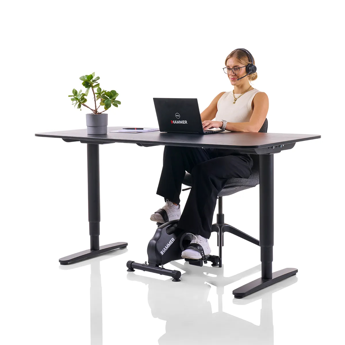 Frau arbeitet am Schreibtisch, benutzt Laptop und Heimtrainer. Pflanze und Headset vorhanden.
