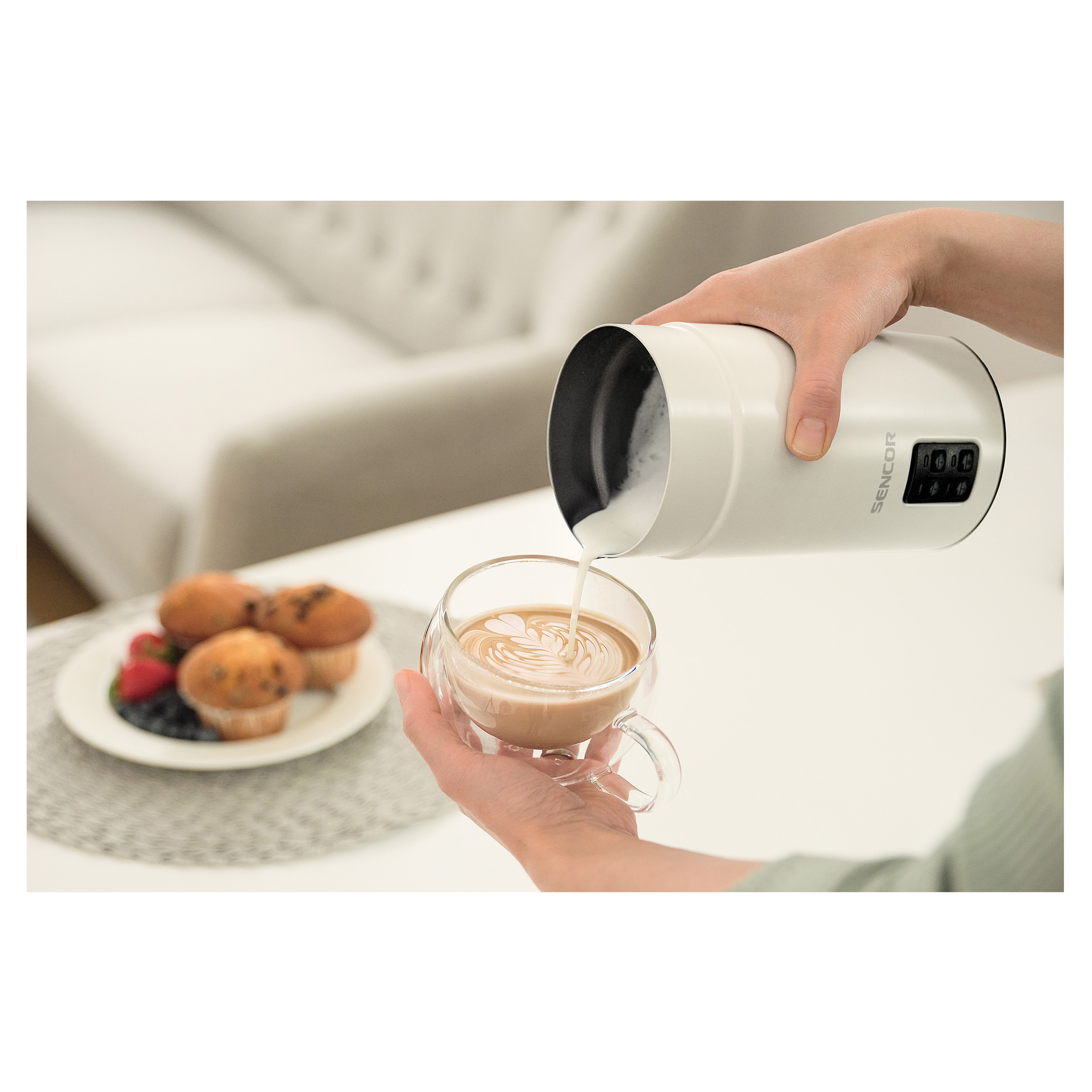 Eine Person gießt Milch aus einem weißen Milchaufschäumer in eine klare Glastasse mit Kaffee.