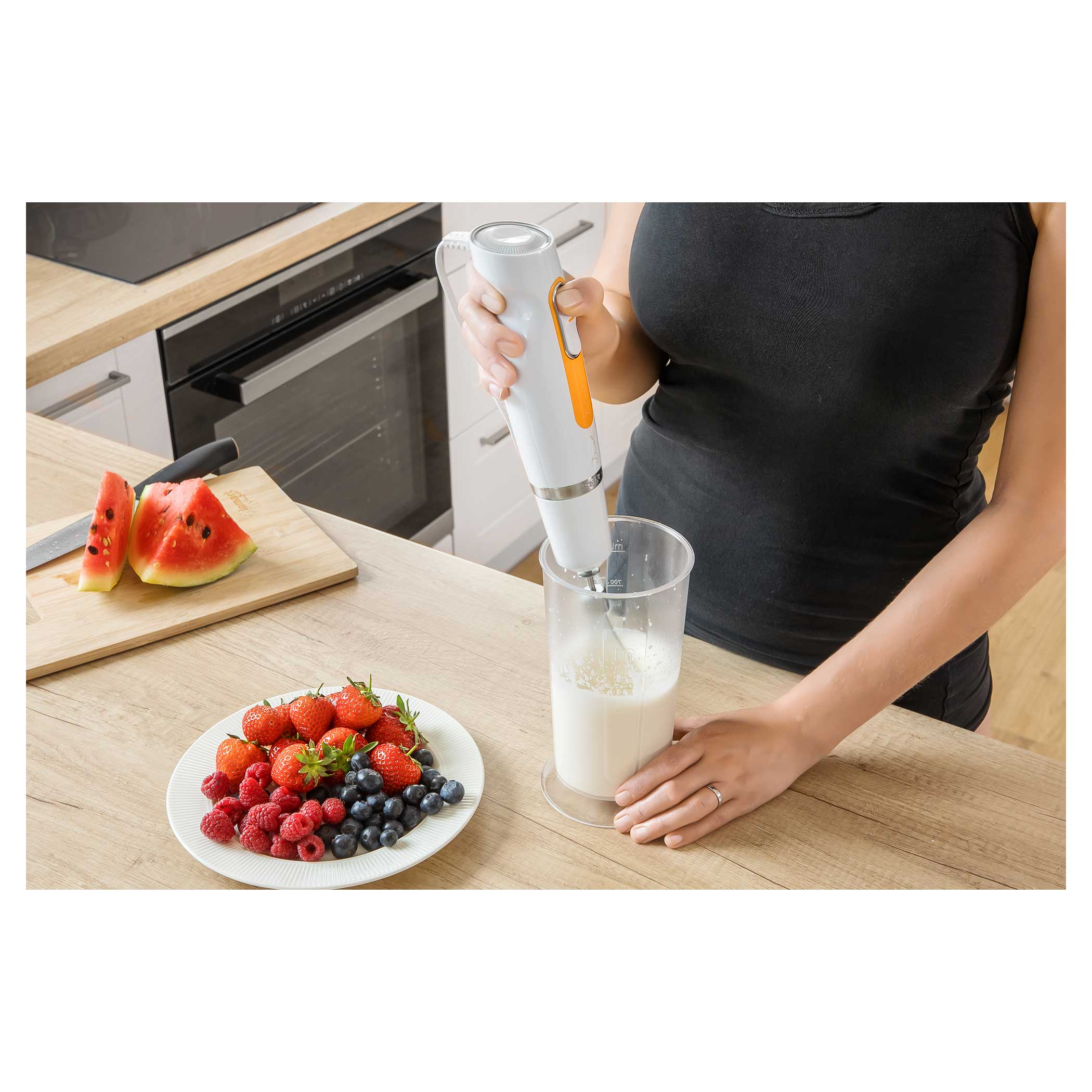 Una persona usa un frullatore a immersione bianco per mescolare ingredienti in un bicchiere trasparente.